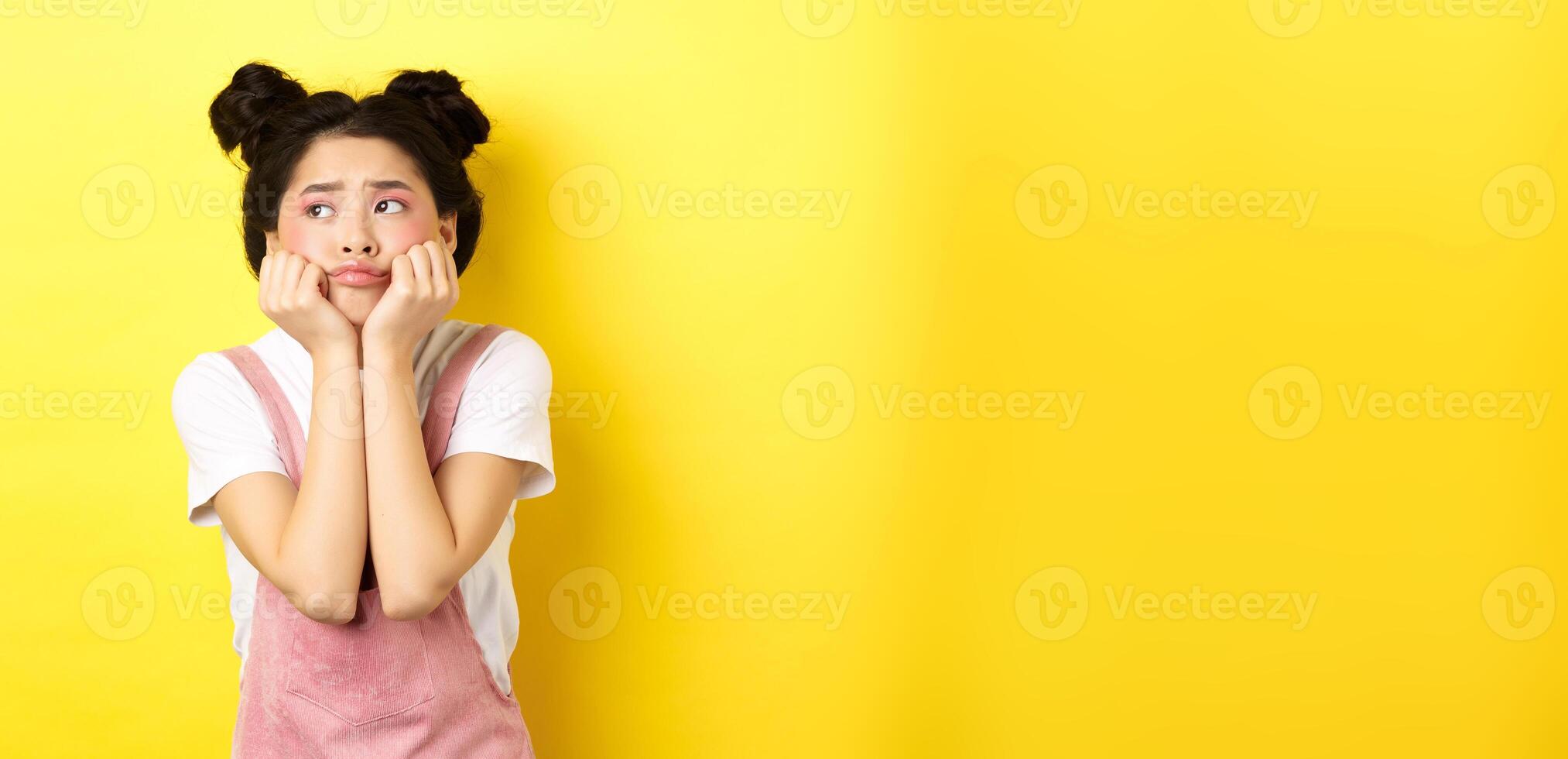 Sad moody asian girl, lean face on hands, looking left with sulking expression, standing in summer clothes on yellow background photo