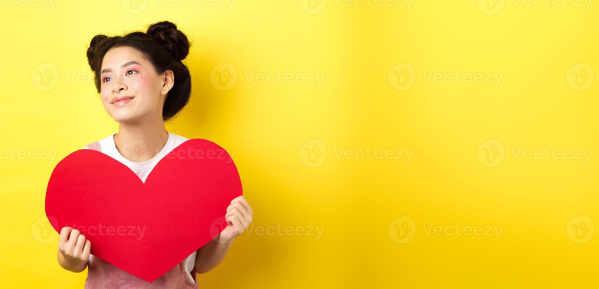Asian girl in love, looking dreamy left and smiling, showing Valentines red heart cutout, imaging romantic date, standing on yellow background photo