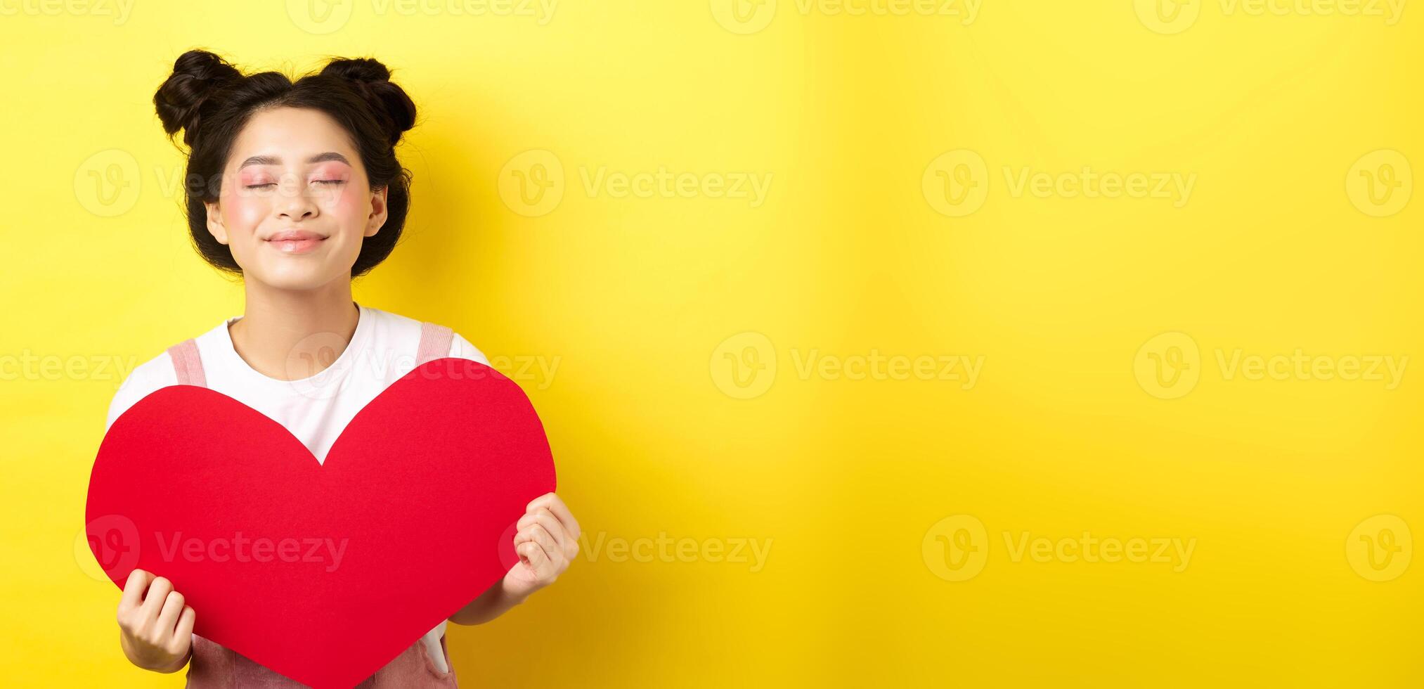 Cute teenage asian girl dreaming of true love, close eyes and showing big red heart cutout, waiting for soulmate on Valentines day, standing with romantic makeup on yellow background photo
