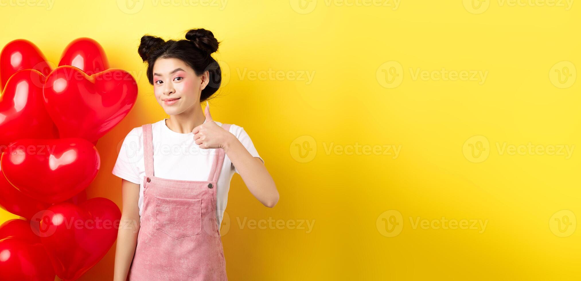 Fashionble asian woman in romantic outfit with make-up, showing thumb up and smiling, praising Valentines day offer, standing near red heart balloons, yellow background photo