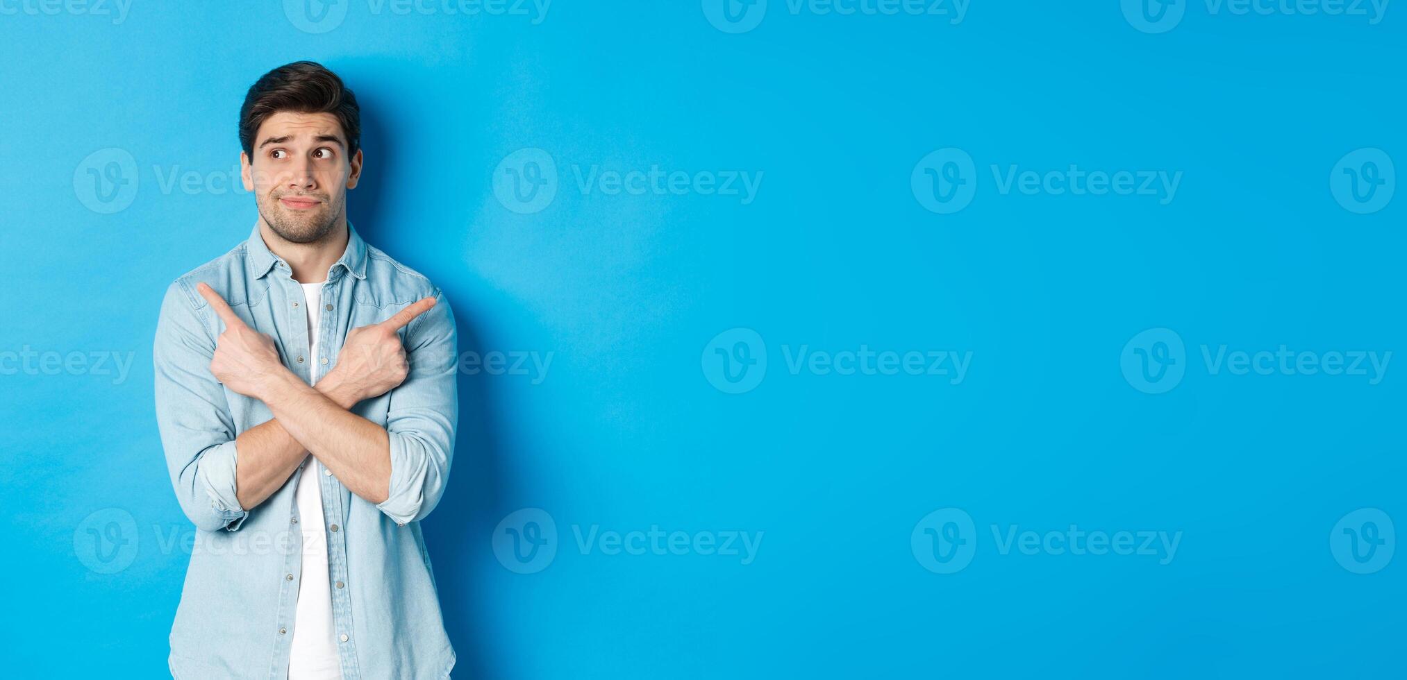 Portrait of indecisive adult man pointing fingers sideways but looking left, making choice between two products, standing against blue background photo