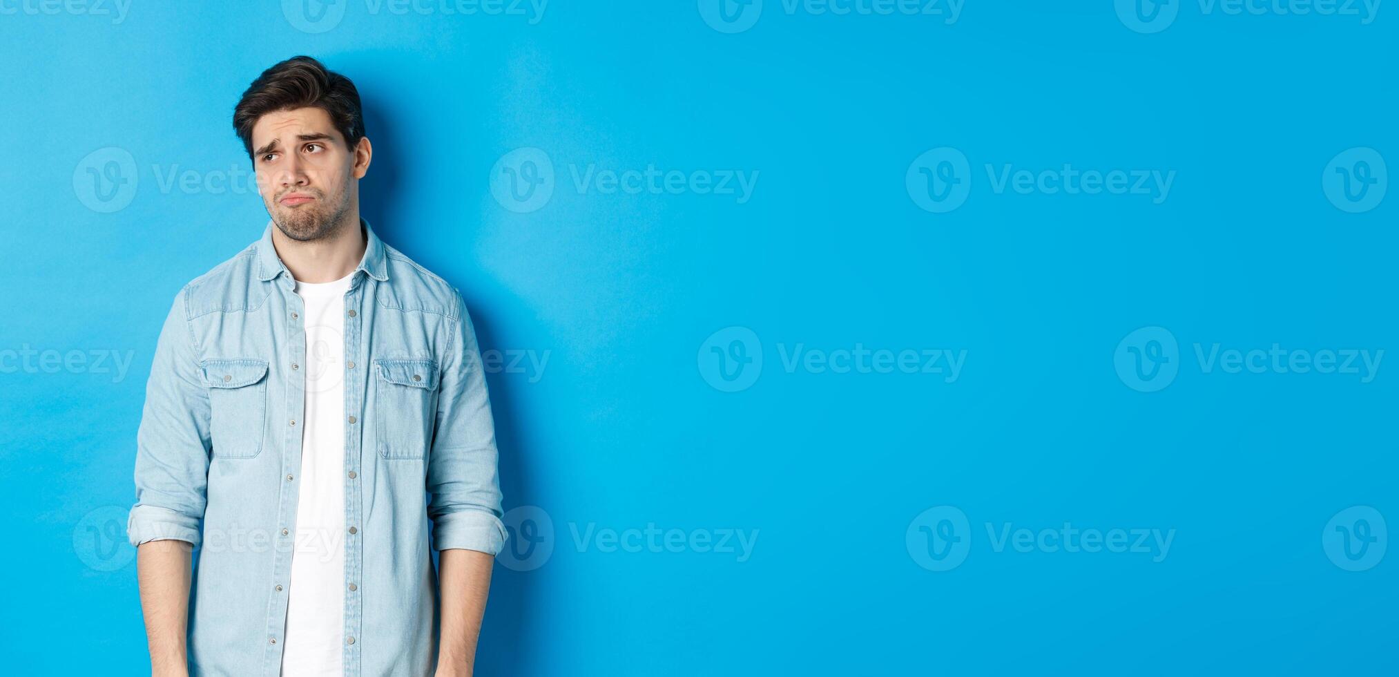 Image of reluctant and sad guy in casual outfit looking left, frowning and feeling upset, standing against blue background photo
