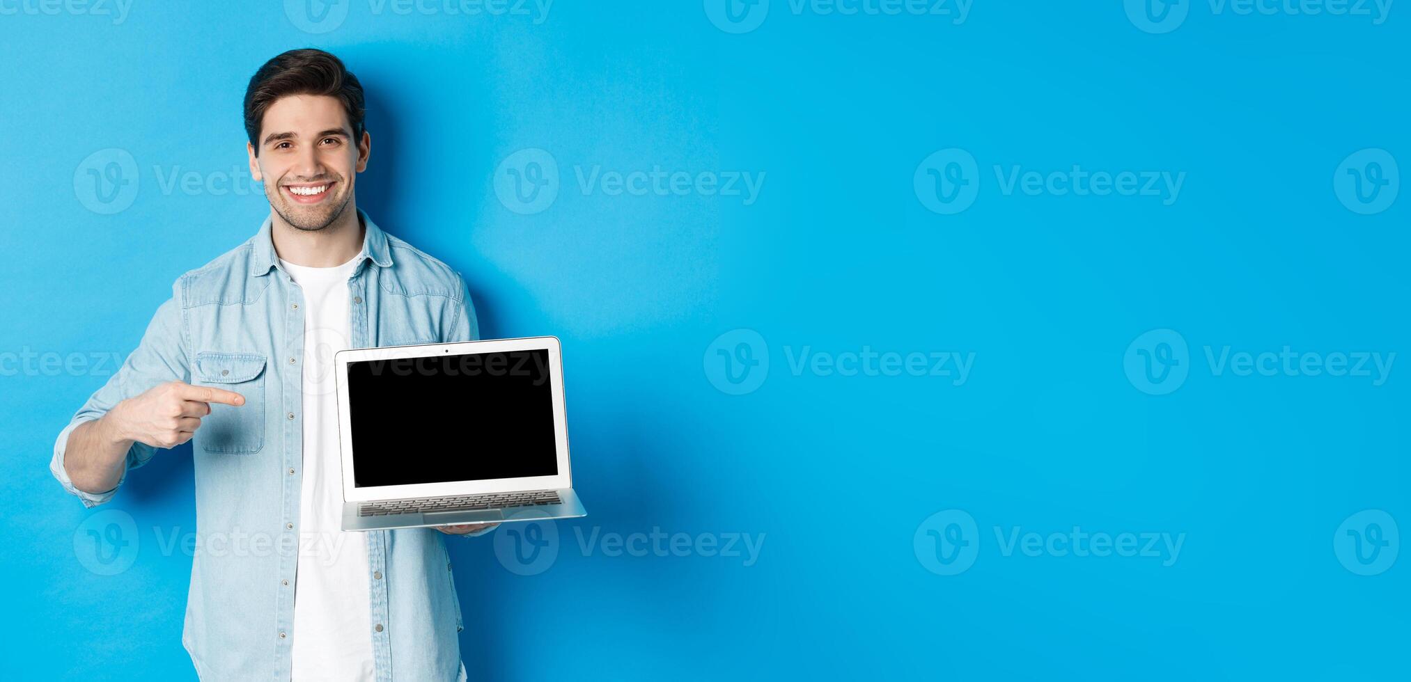 Handsome young man pointing finger at screen of computer, smiling pleased, showing promo in internet or website, standing over blue background photo