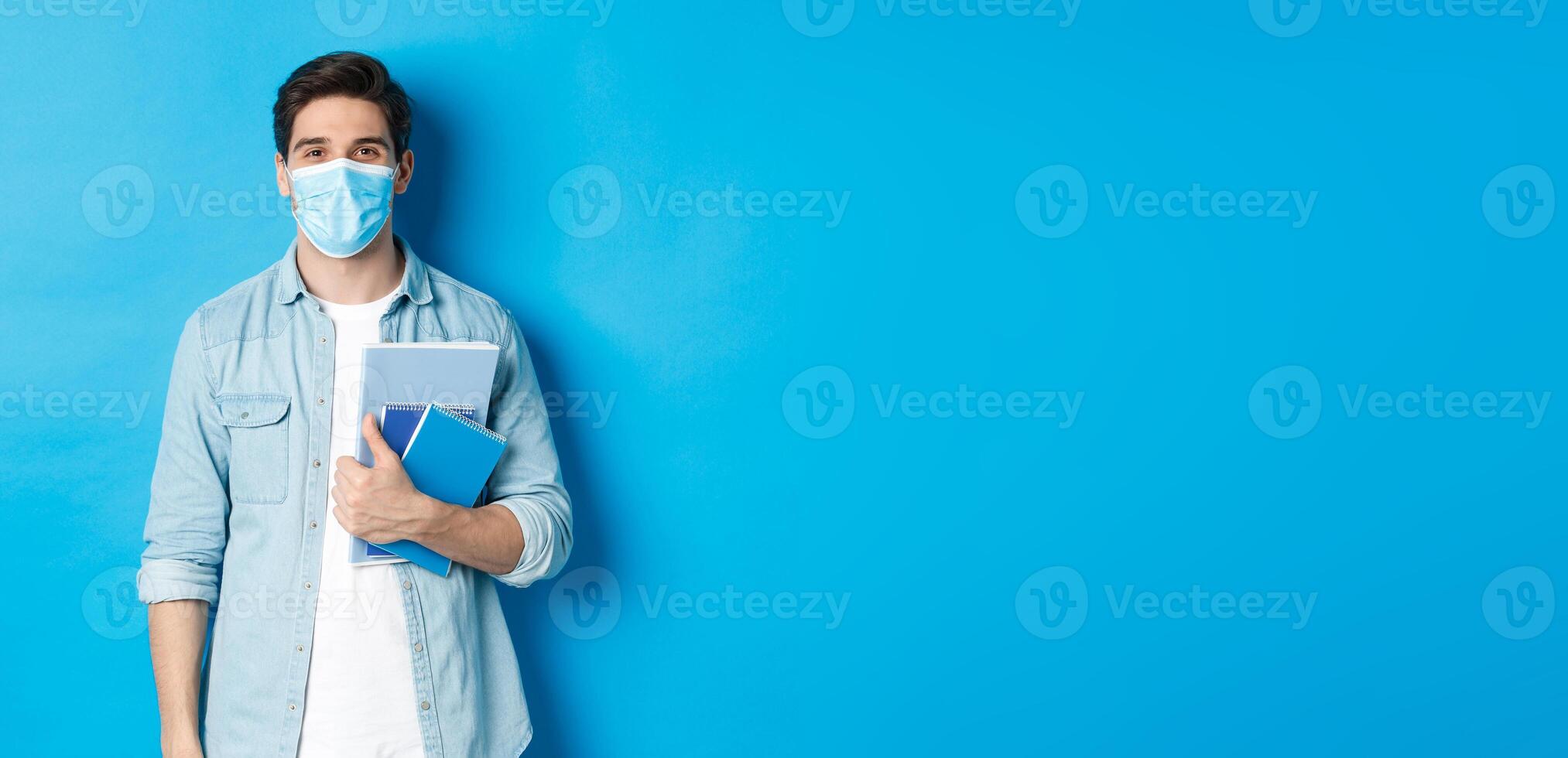 Education, covid-19 and social distancing. Guy student in medical mask looking happy, holding notebooks, standing over blue background photo