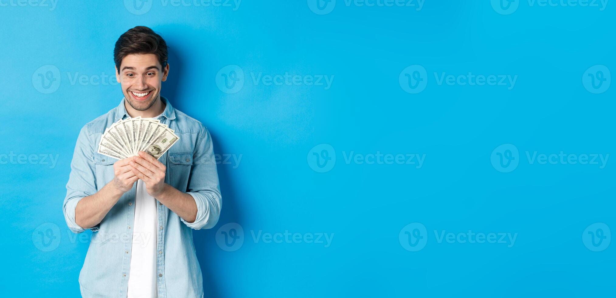Excited successful man counting money, looking satisfied at cash and smiling, standing over blue background photo