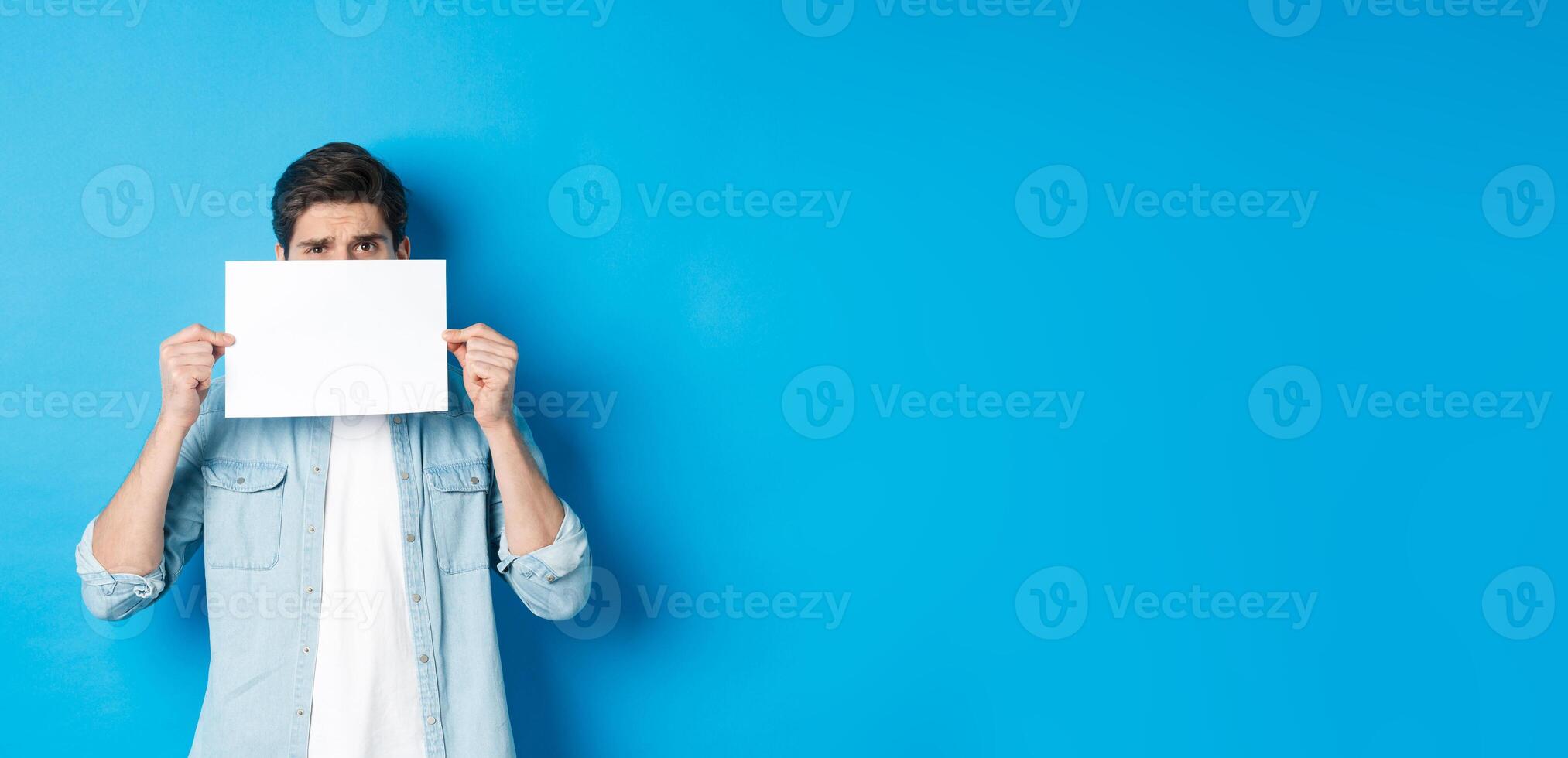 Shy and nervous guy hiding face behind blank piece of paper, peeking with worried look, showing your logo, standing over blue background photo