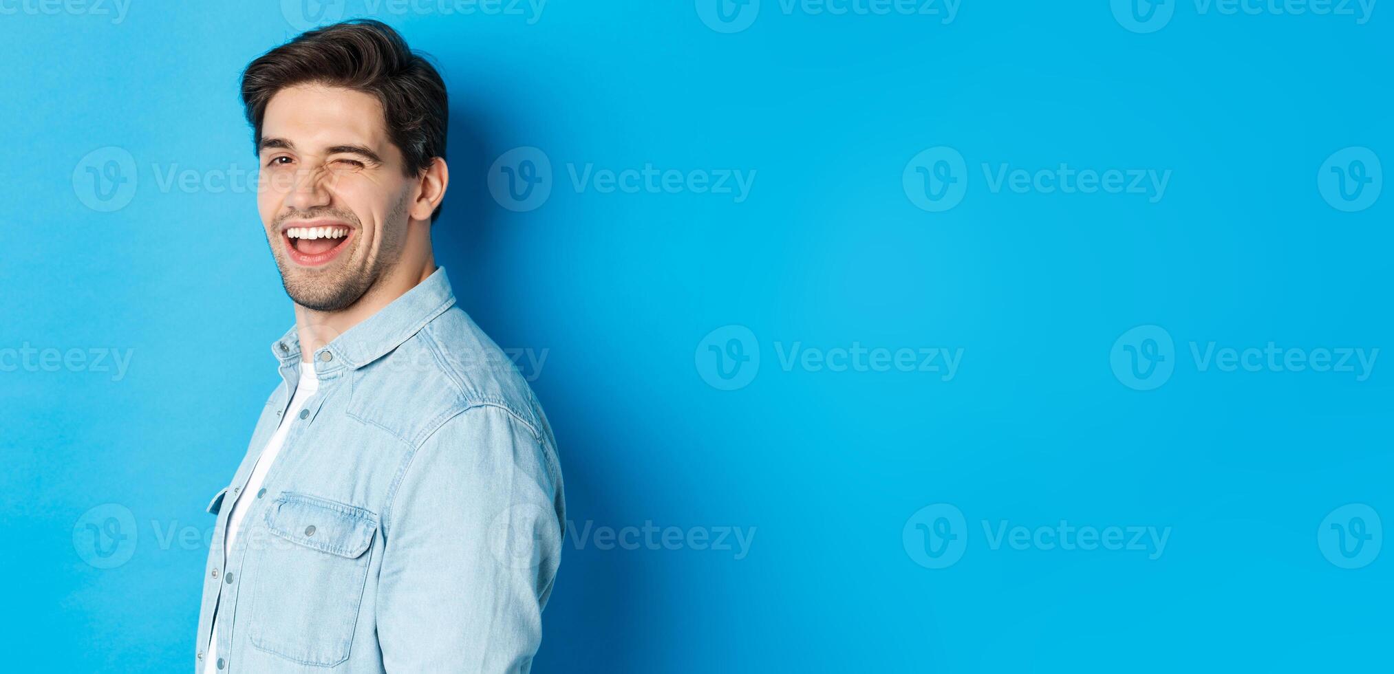Happy and confident man turn head at camera, winking and smiling, standing over blue background photo