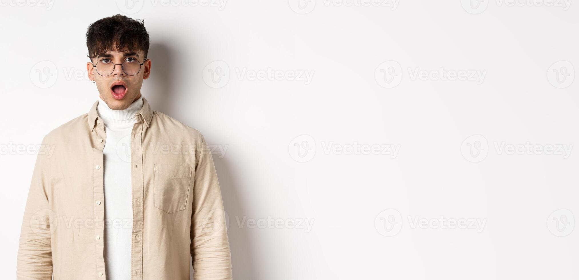 Portrait of shocked young man in glasses drop jaw, open mouth and stare amazed at camera, looking at something impressive, standing on white background photo