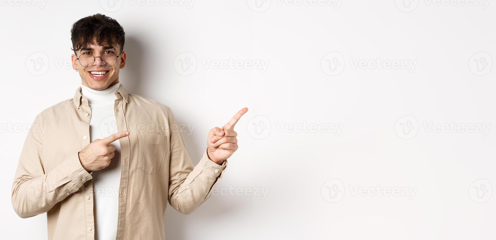 Proud and happy young man in glasses showing logo, pointing fingers at upper right corner and smiling, standing on white background photo