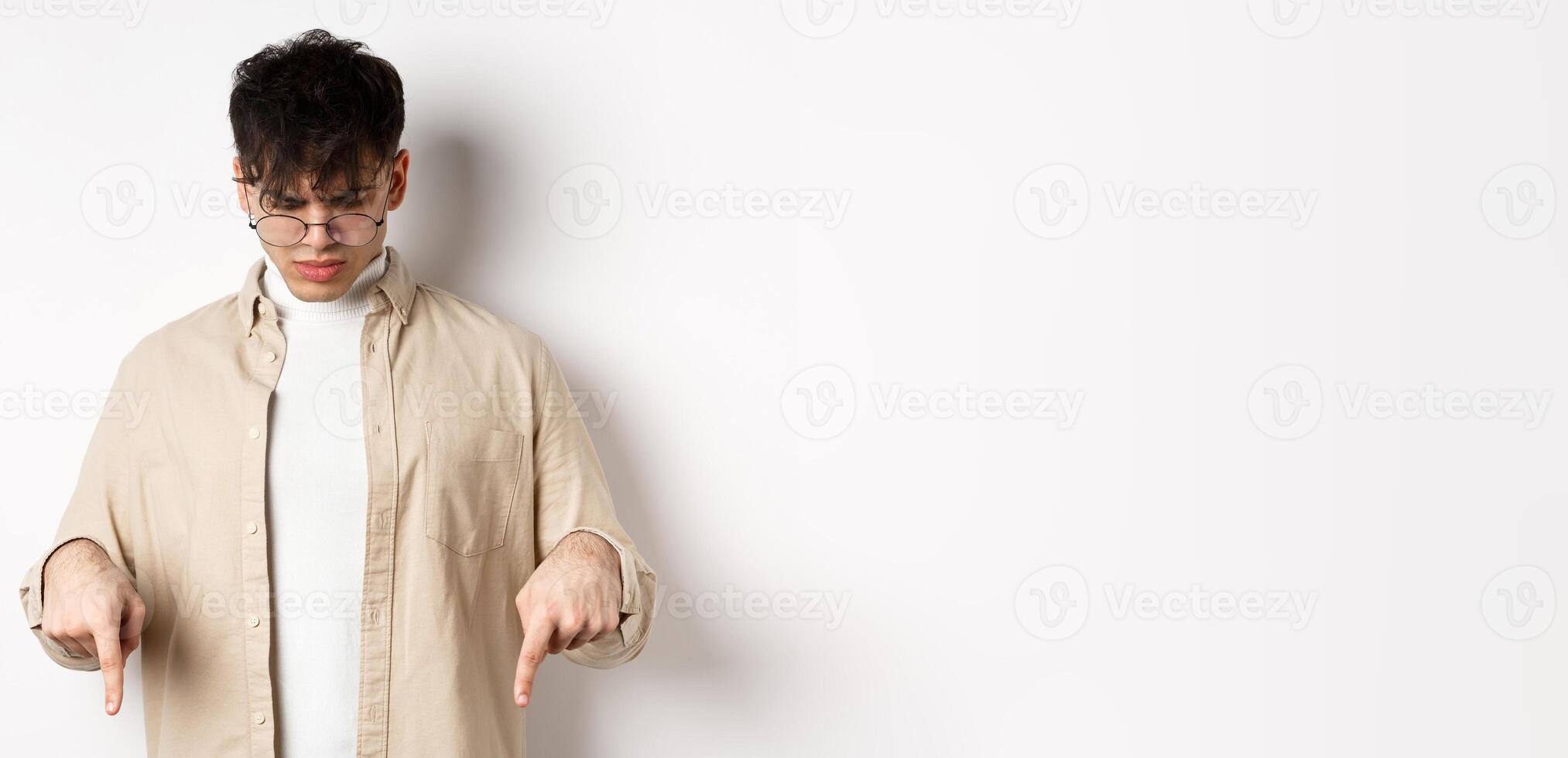 Confused frowning guy in glasses looking, pointing fingers down at something strange, standing puzzled on white background photo