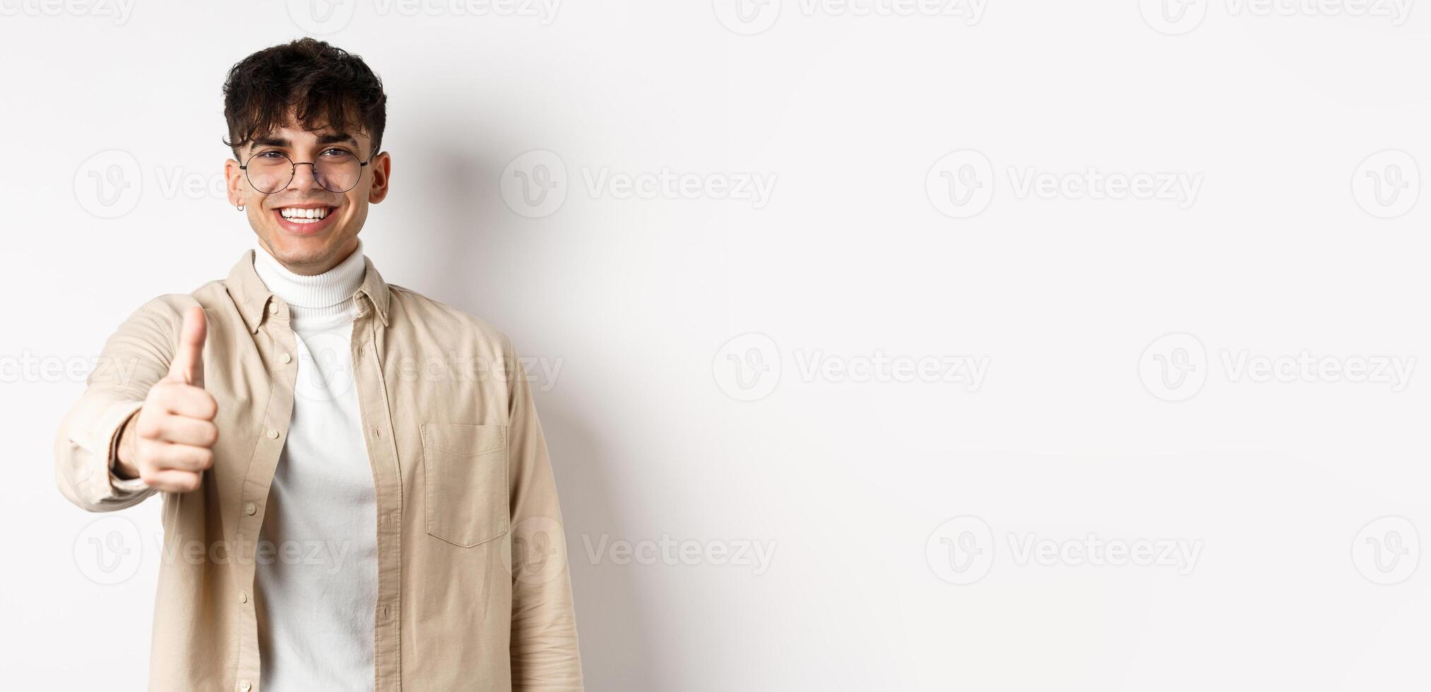 Image of happy young man showing thumb up and smiling satisfied, praise good job, say well done and look pleased, standing on white background photo