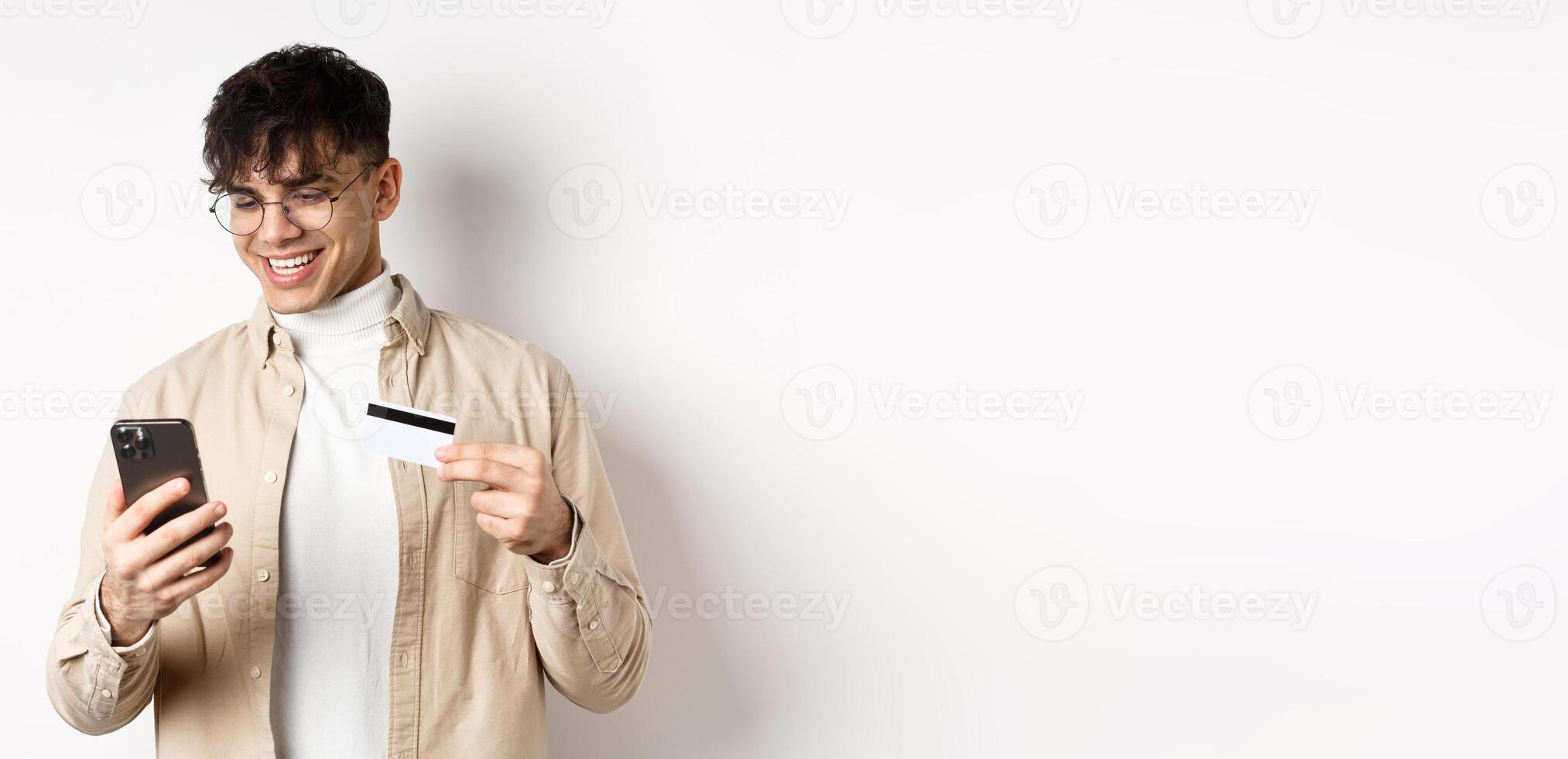 Online shopping. Happy young man using smartphone and plastic credit card, paying in internet, standing on white background photo