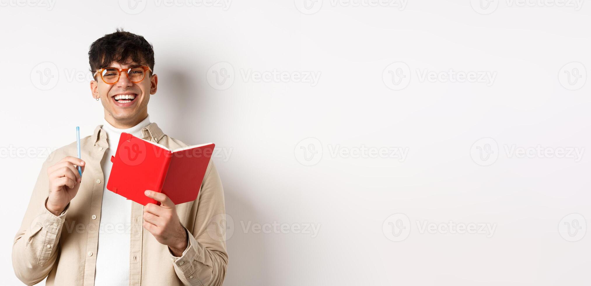 Cheerful young man in glasses laughing and taking notes, writing down in planner, holding pen and diary, standing on white background photo