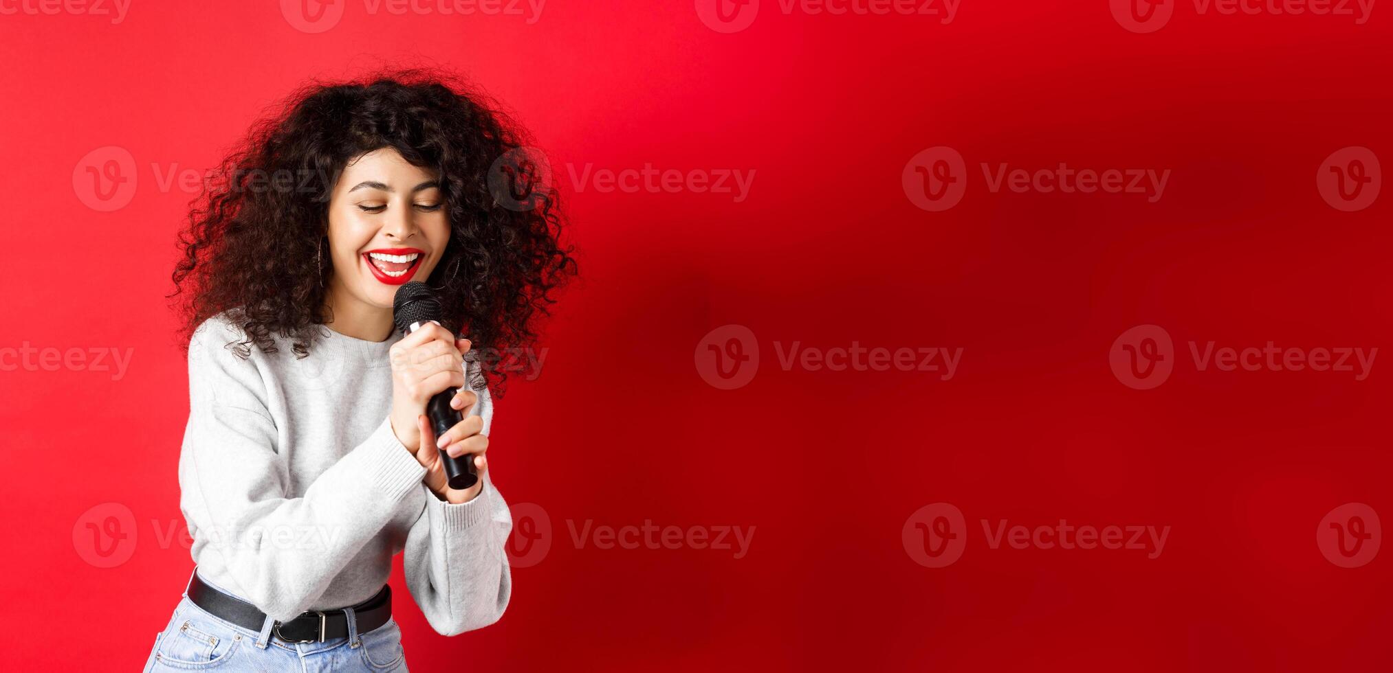 Hobbies and leisure concept. Beautiful woman singing in microphone and smiling, having fun at karaoke, standing on red background photo