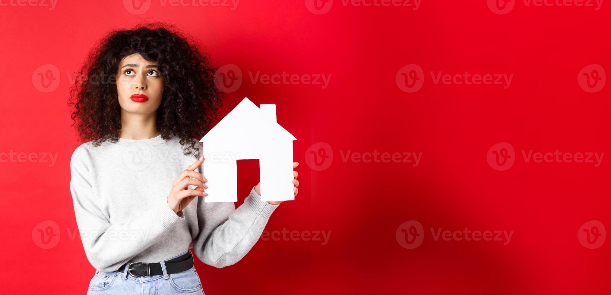 Real estate. Sad woman thinking of buying own house, showing paper home cutout and looking up with uspet face, standing on red background photo