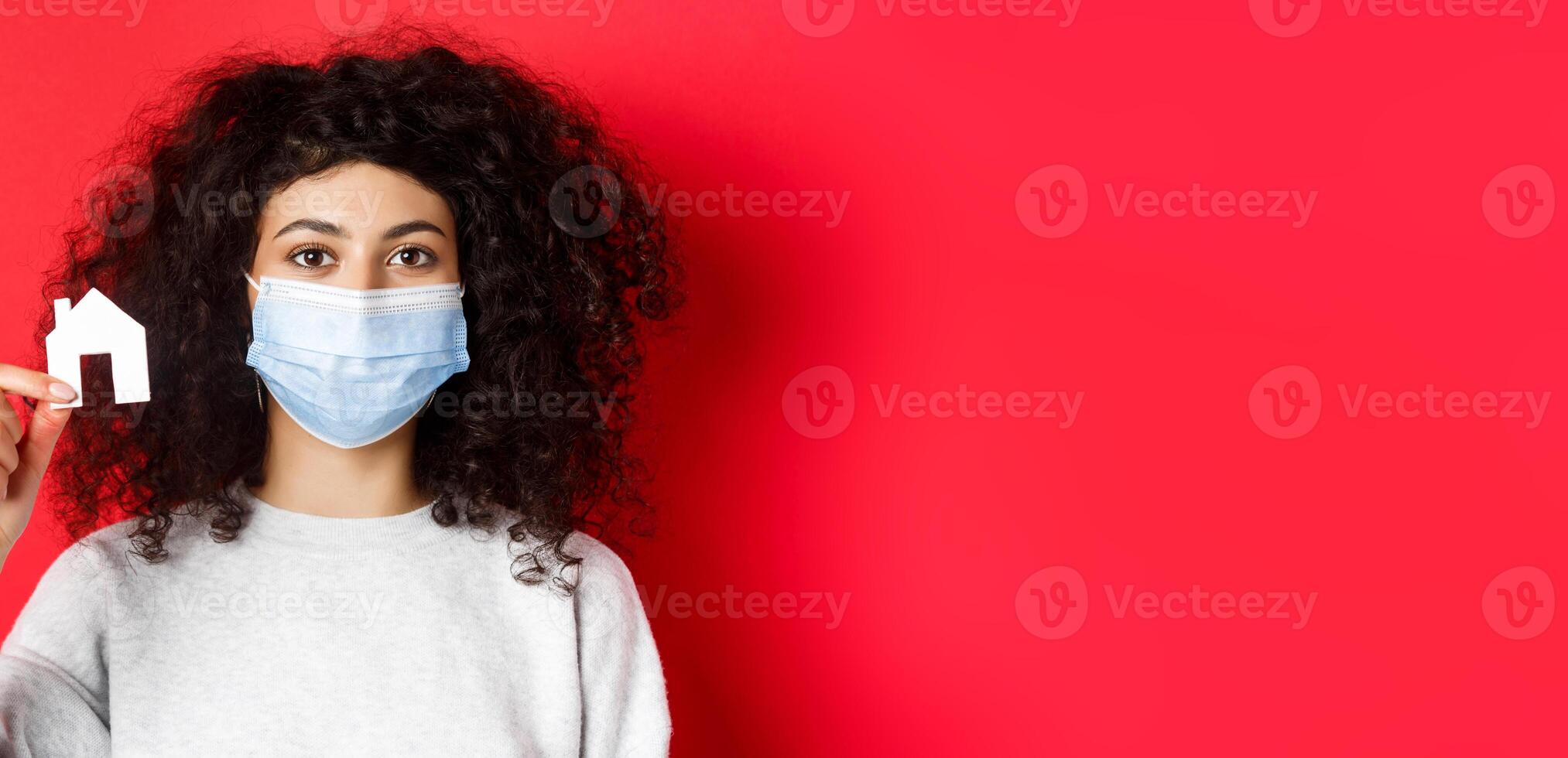 Real estate and covid-19 concept. Excited woman in medical mask showing small paper house cutout, standing on red background photo