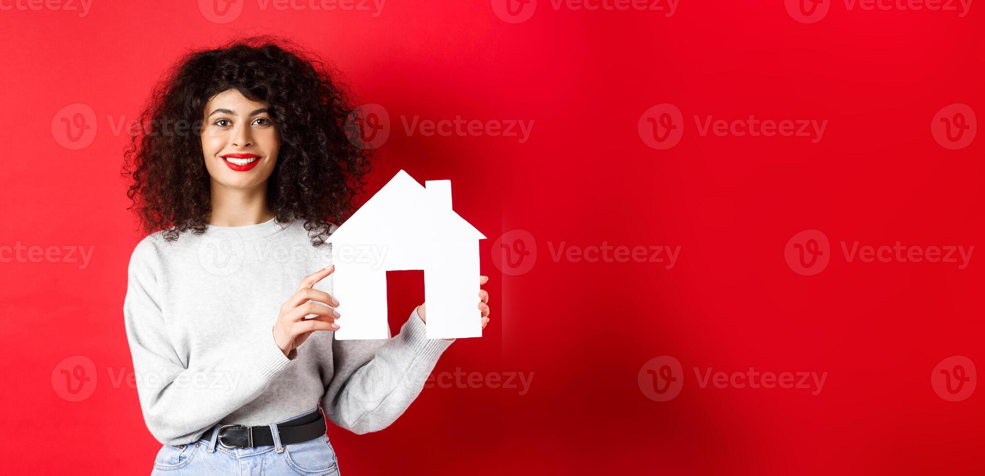 Real estate. Smiling caucasian woman with curly hair and red lips, showing paper house model, searching property, standing on red background photo