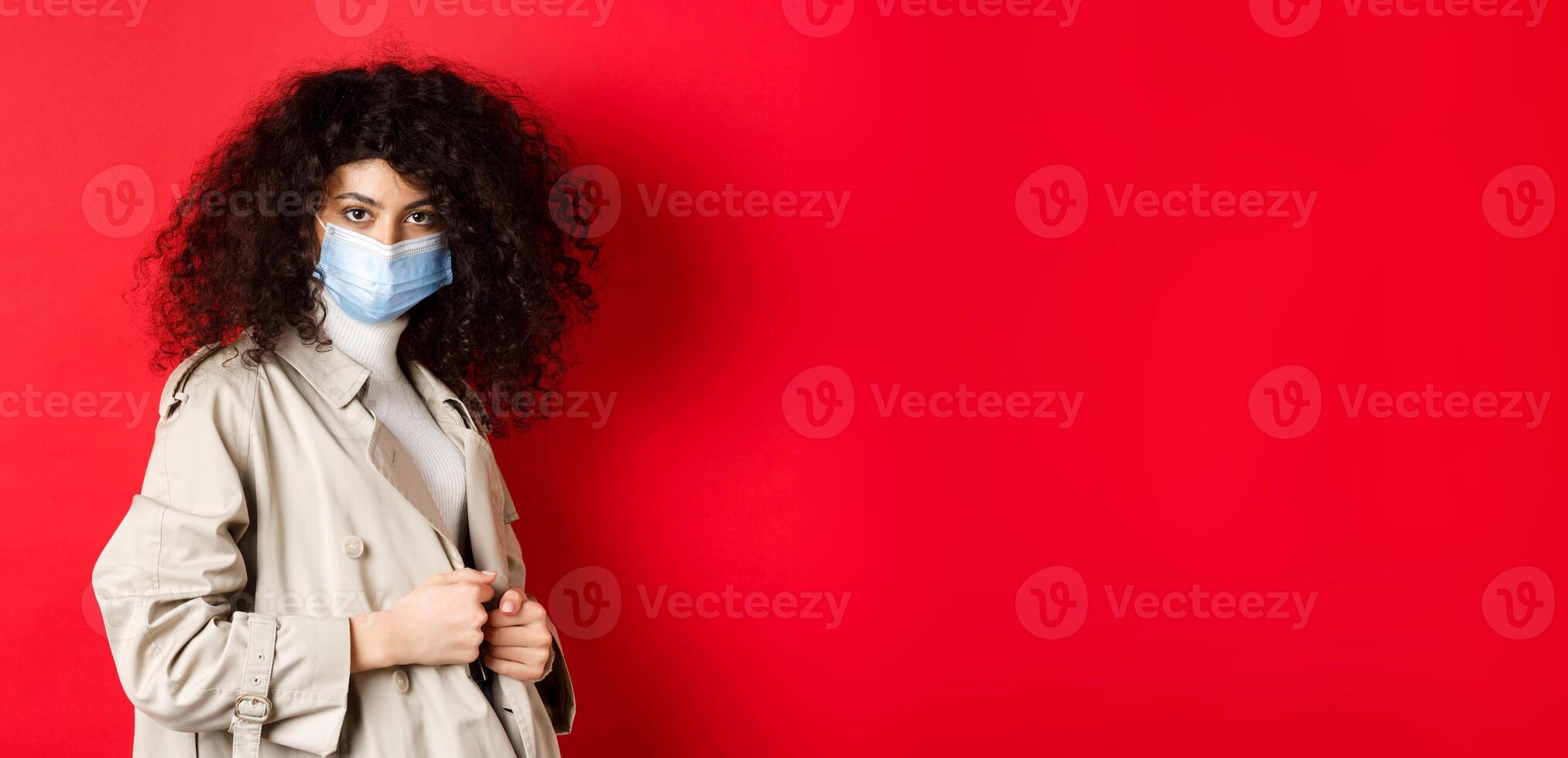 Covid-19, pandemic and quarantine concept. Sassy girl in trench coat and medical mask, put on trench coat for spring walk, red background photo