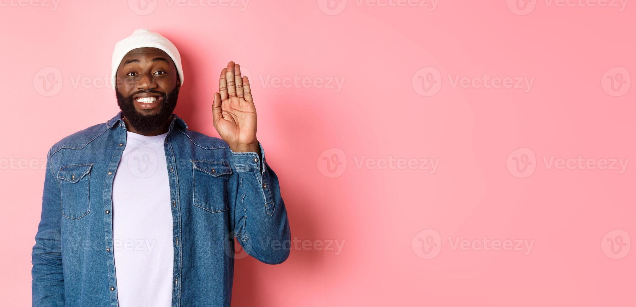Friendly smiling Black man saying hello, waving hand, greeting you, standing over pink background photo