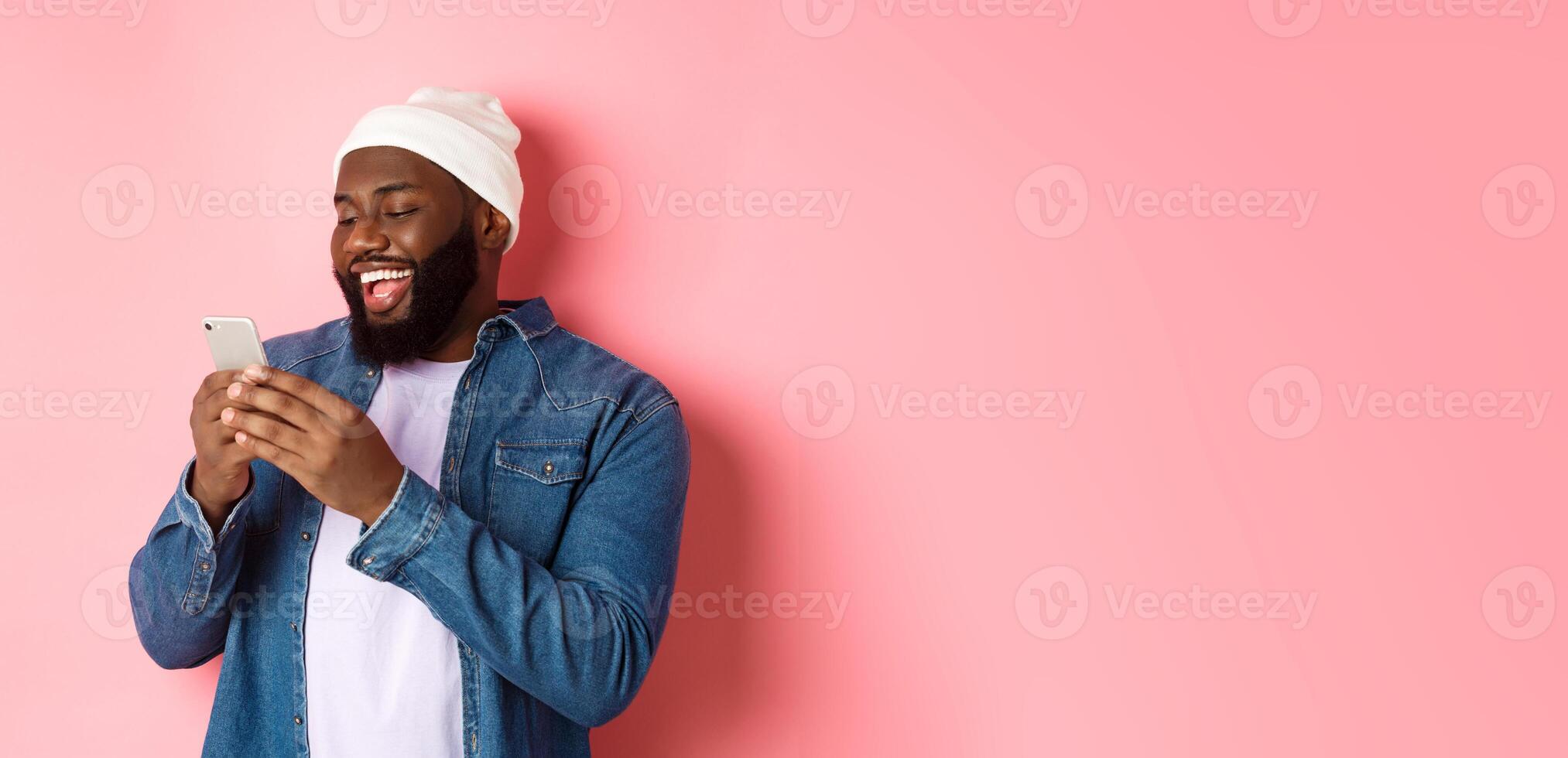 Technology and online shopping concept. Happy Black bearded man reading message and smiling, using smartphone against pink background photo