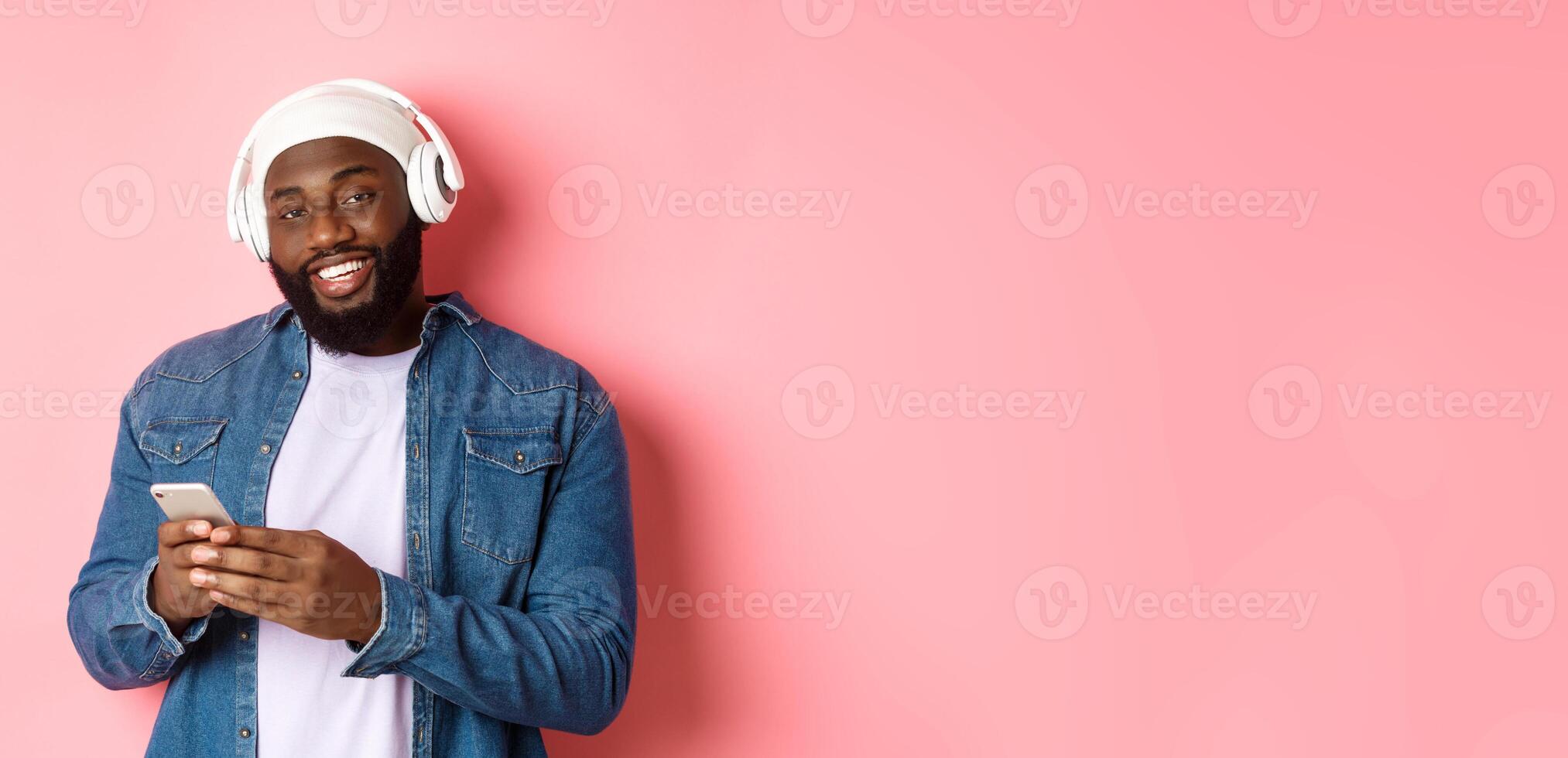Handsome hipster guy in headphones smiling satisfied at camera, listening music in headphones, using mobile app, standing over pink background photo