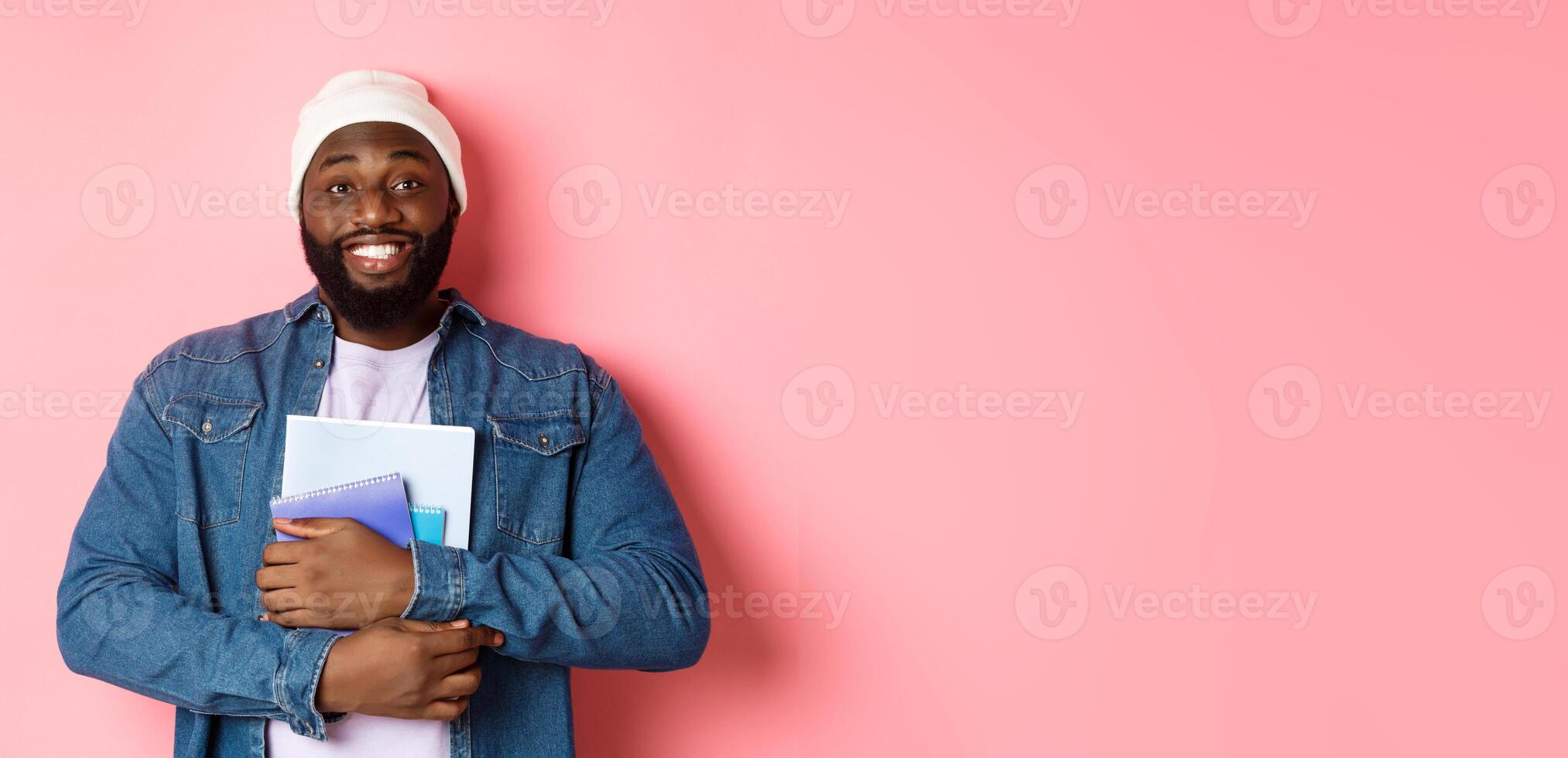 Image of adult Black man holding notebooks and smiling, studying at courses, standing over pink background photo