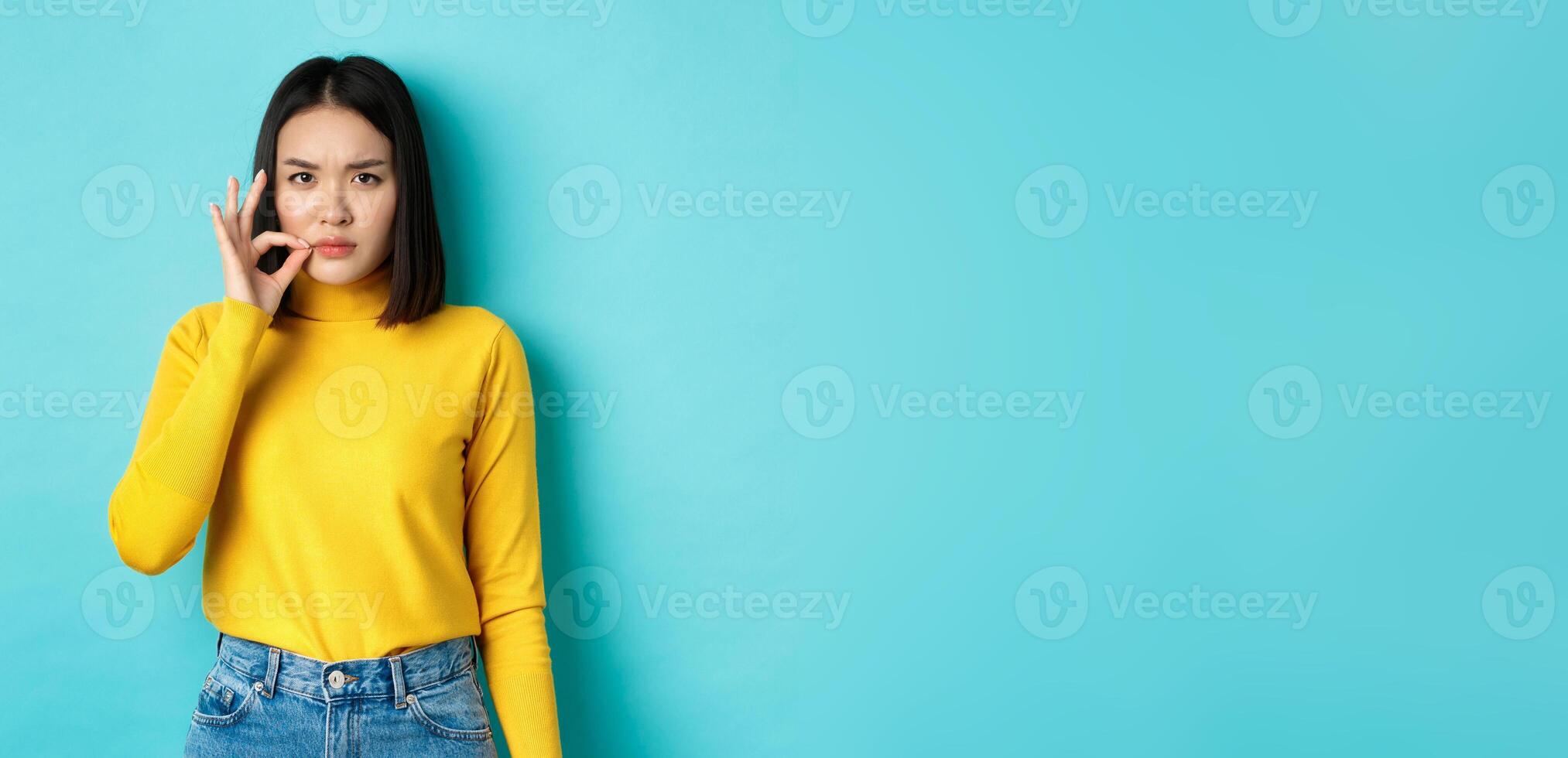 Serious asian girl showing mouth zip gesture, promise to keep shut and frowning, telling big secret, standing in yellow pullover over blue background photo