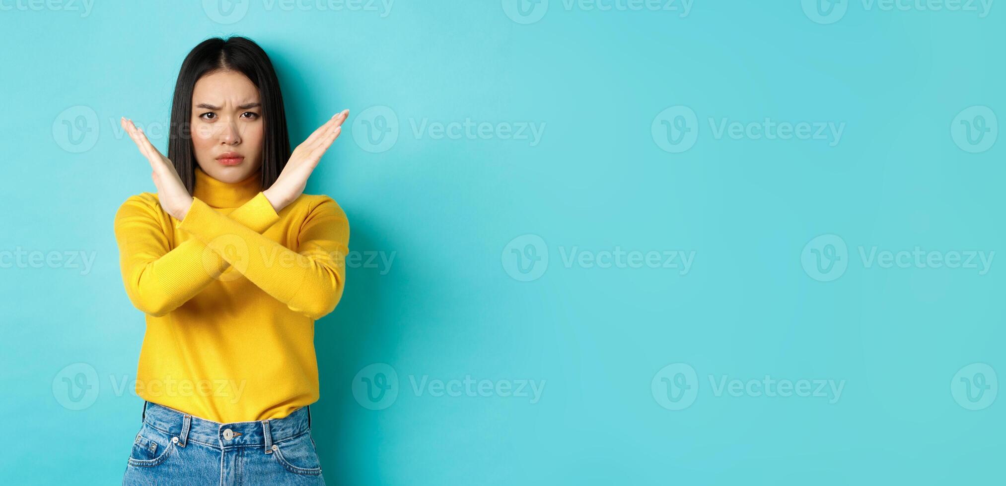 Serious and assertive korean woman showing cross stop gesture, frowning and telling no, prohibit action, disapprove something bad, standing over blue background photo