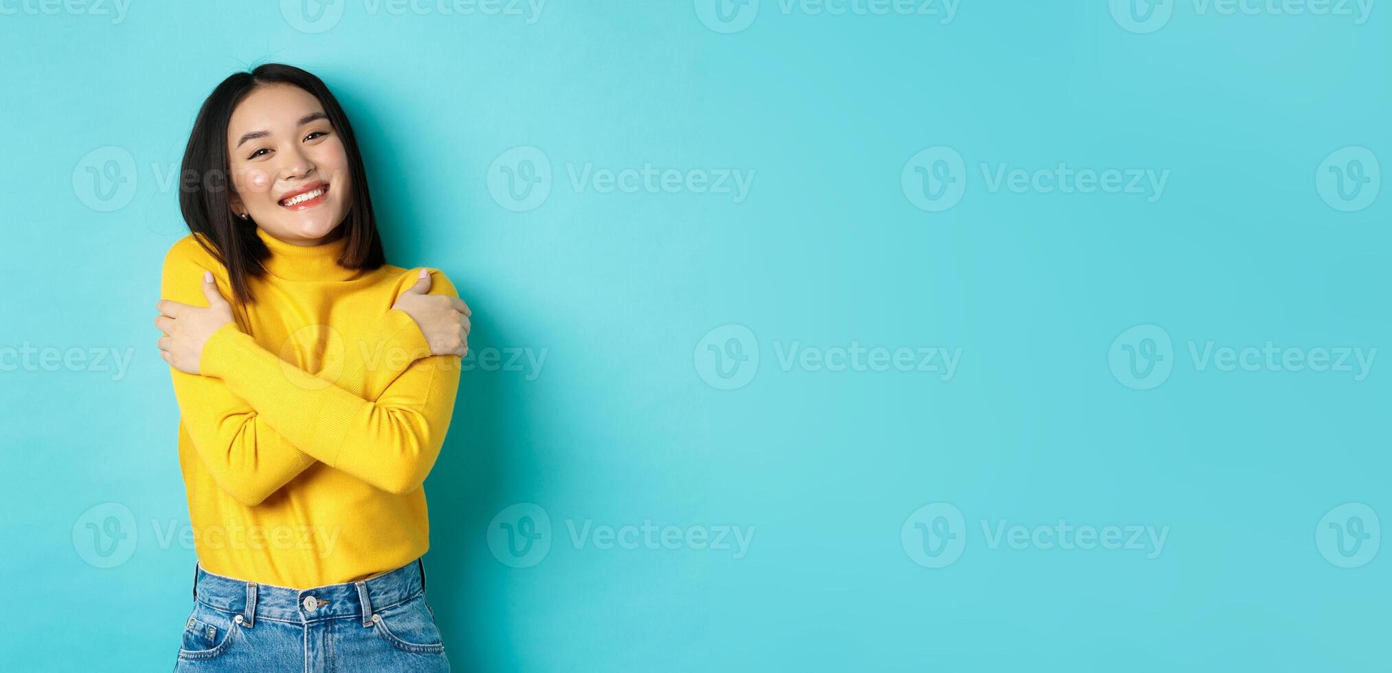 Cheerful beautiful girl embracing own body, smiling and cuddling herself, standing carefree in yellow pullover against blue background photo