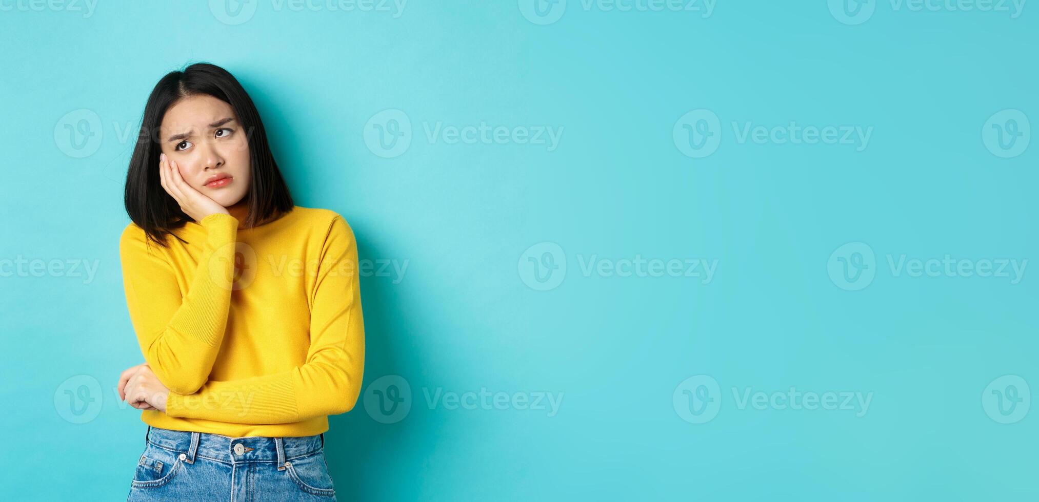 Sad and bored asian teenage girl leaning on palm, looking at upper left corner with lonely and upset expression, standing over blue background photo