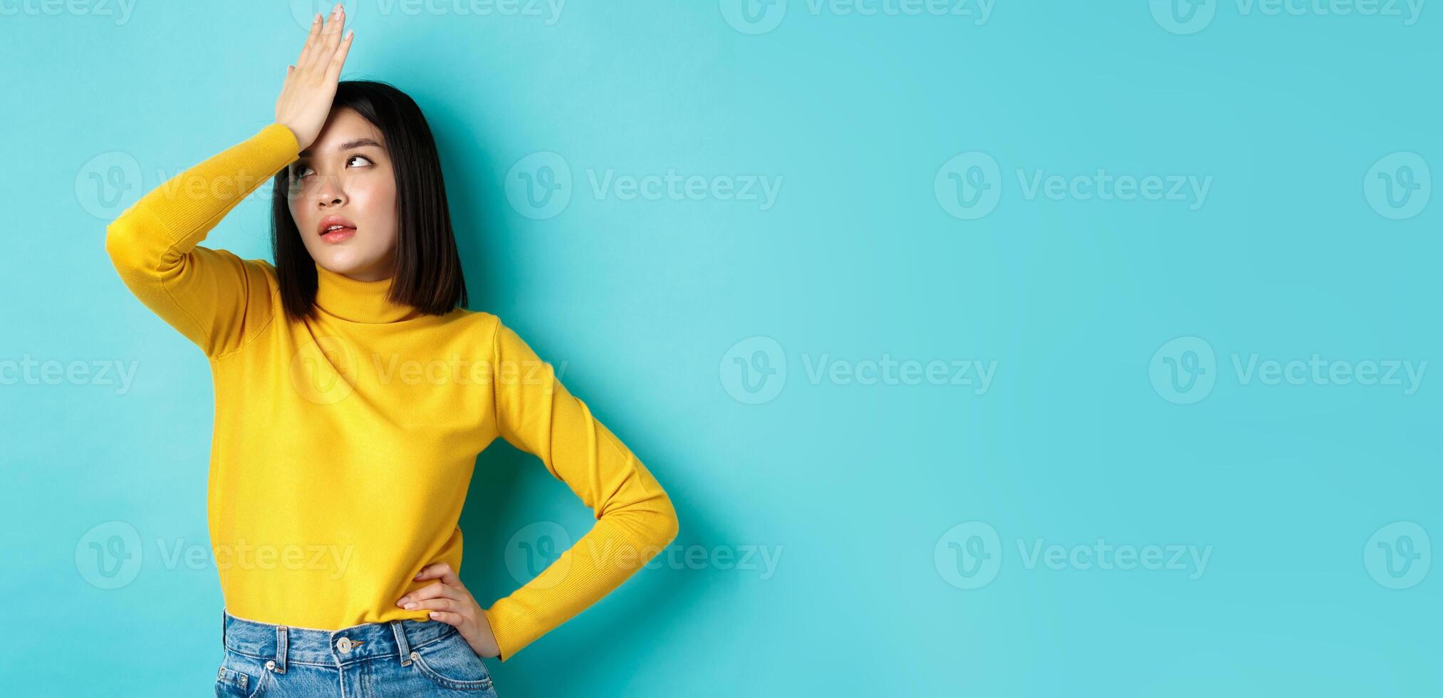 Emotions and lifestyle concept. Annoyed asian girl roll eyes and face palm, standing bothered in yellow pullover against blue background photo