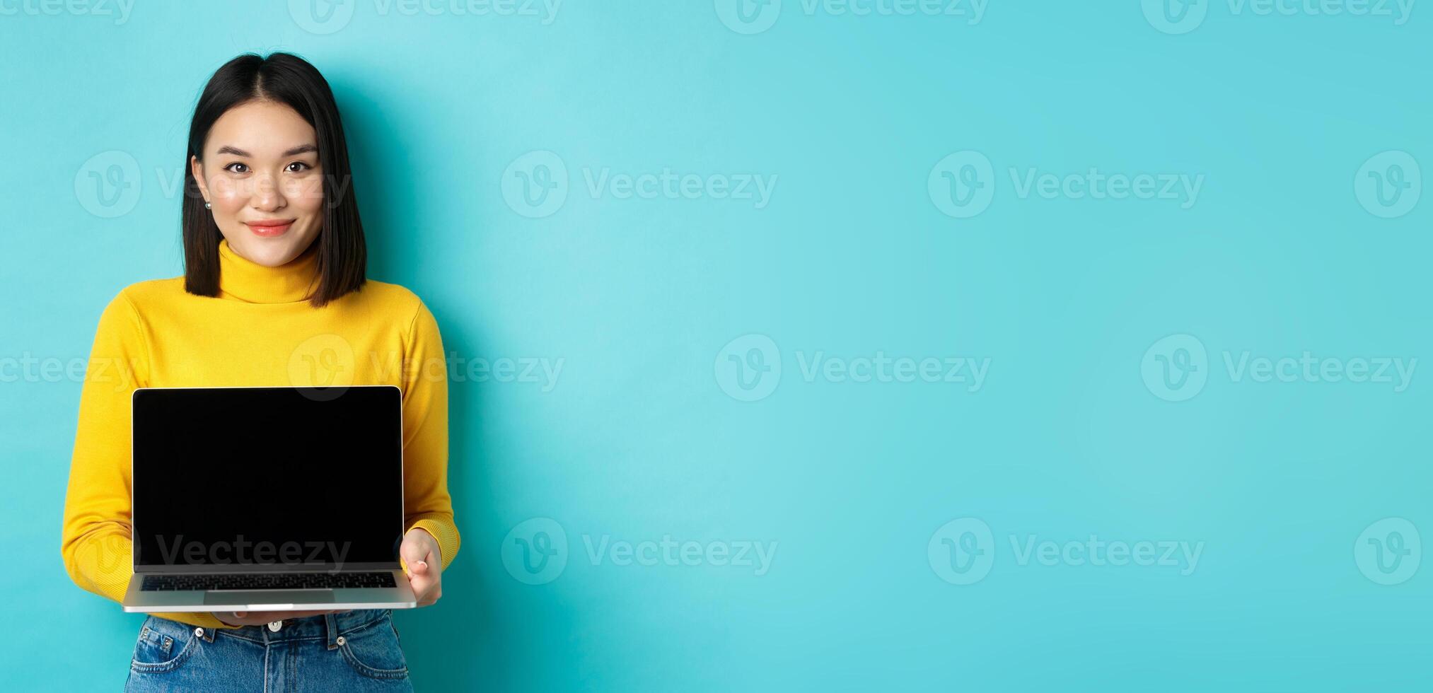 Beautiful and stylish asian woman demonstrate product on screen, showing empty laptop display and smiling, standing over blue background photo