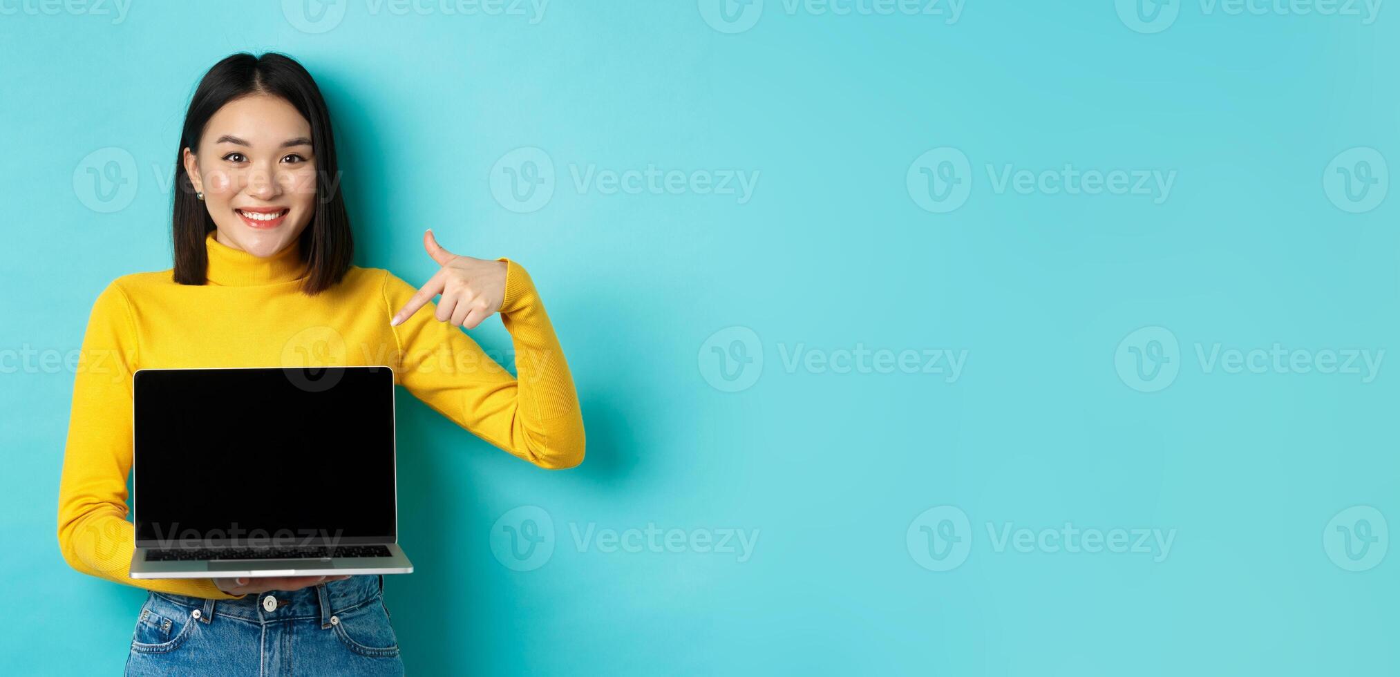 E-commerce and online shopping concept. Beautiful young woman pointing and showing laptop screen, smiling at camera, standing over blue background photo