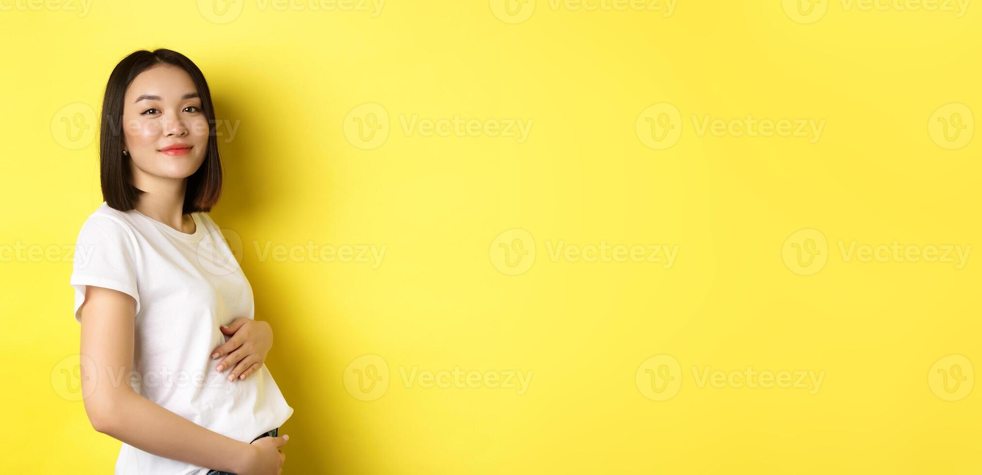 Asian woman smiling kindly, touching belly and looking at camera, being pregnant, having a baby, standing over yellow background photo
