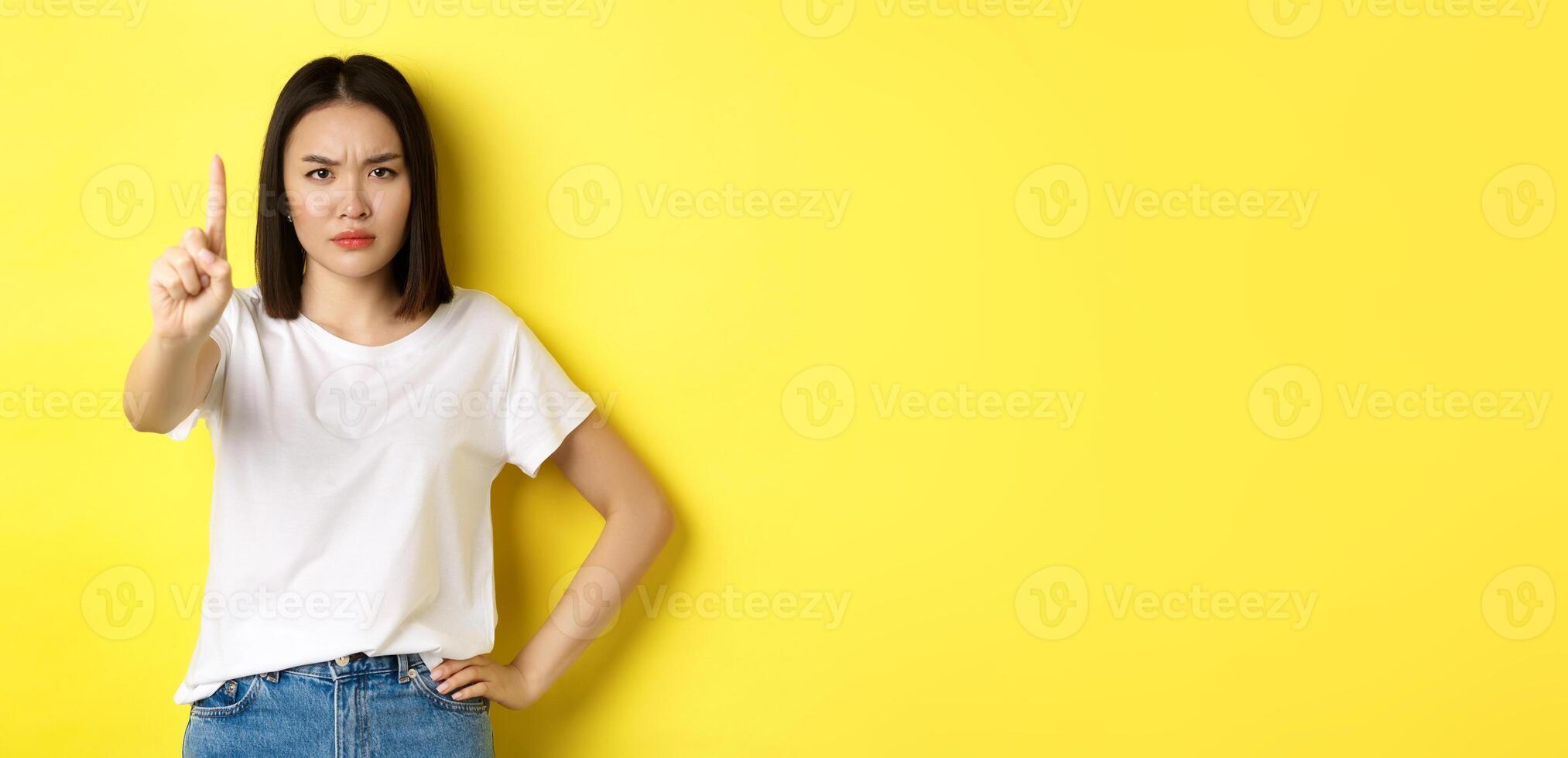 Confident and serious woman tell no, showing extended finger to stop and prohibit something bad, frowning and looking at camera self-assured, standing over yellow background photo
