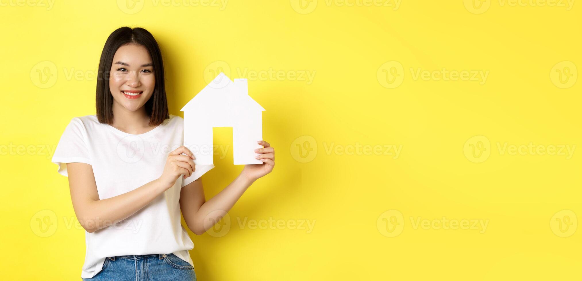 Real estate concept. Smiling beautiful woman showing paper house cutout and looking at camera, buying property, standing over yellow background photo