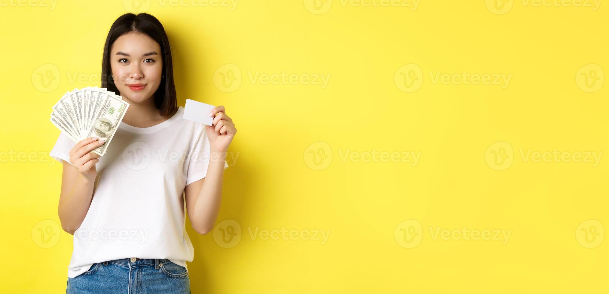 Beautiful asian woman with short dark hair, wearing white t-shirt, showing money in dollars and plastic credit card, standing over yellow background photo