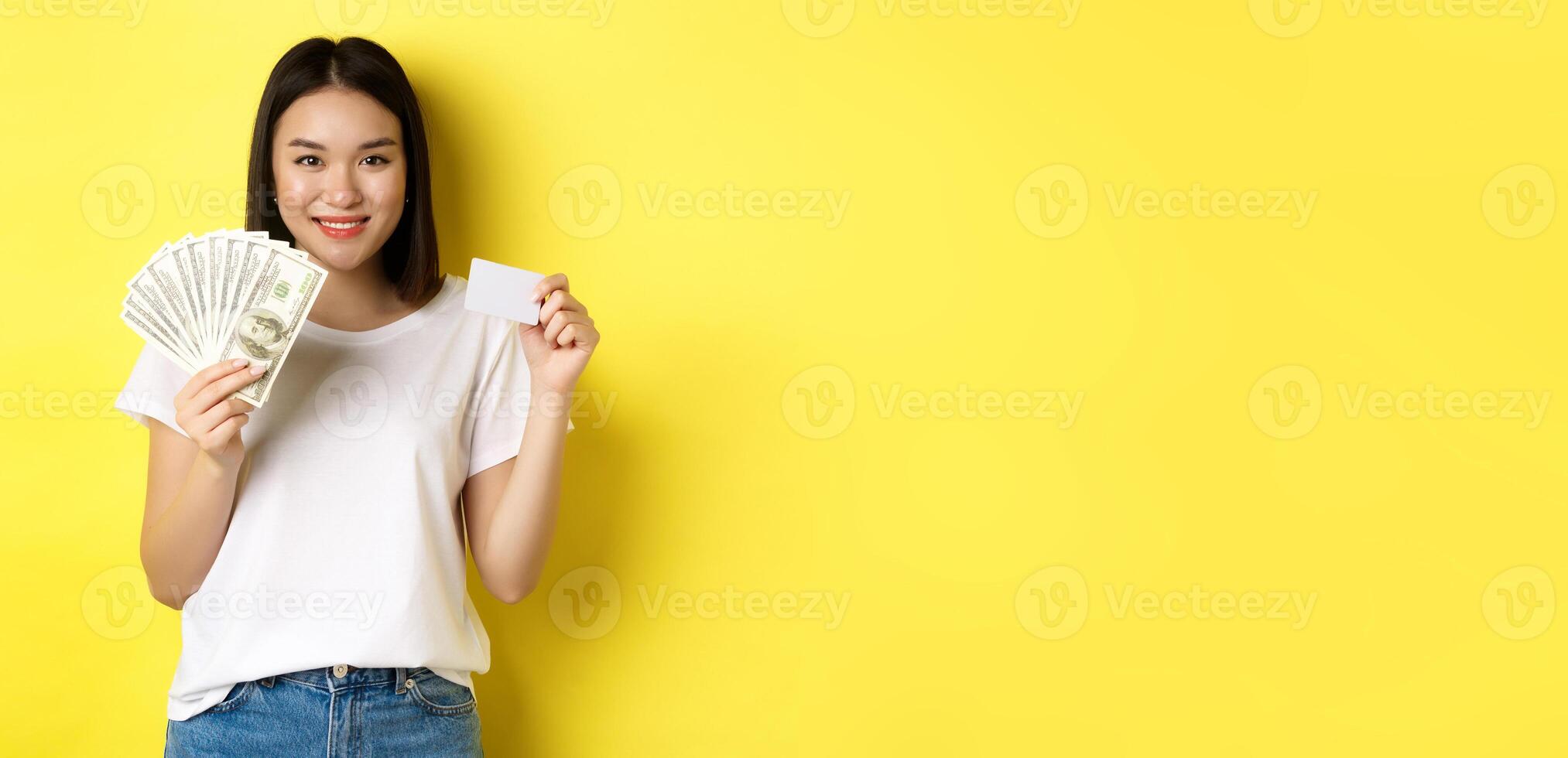 Beautiful asian woman with short dark hair, wearing white t-shirt, showing money in dollars and plastic credit card, standing over yellow background photo