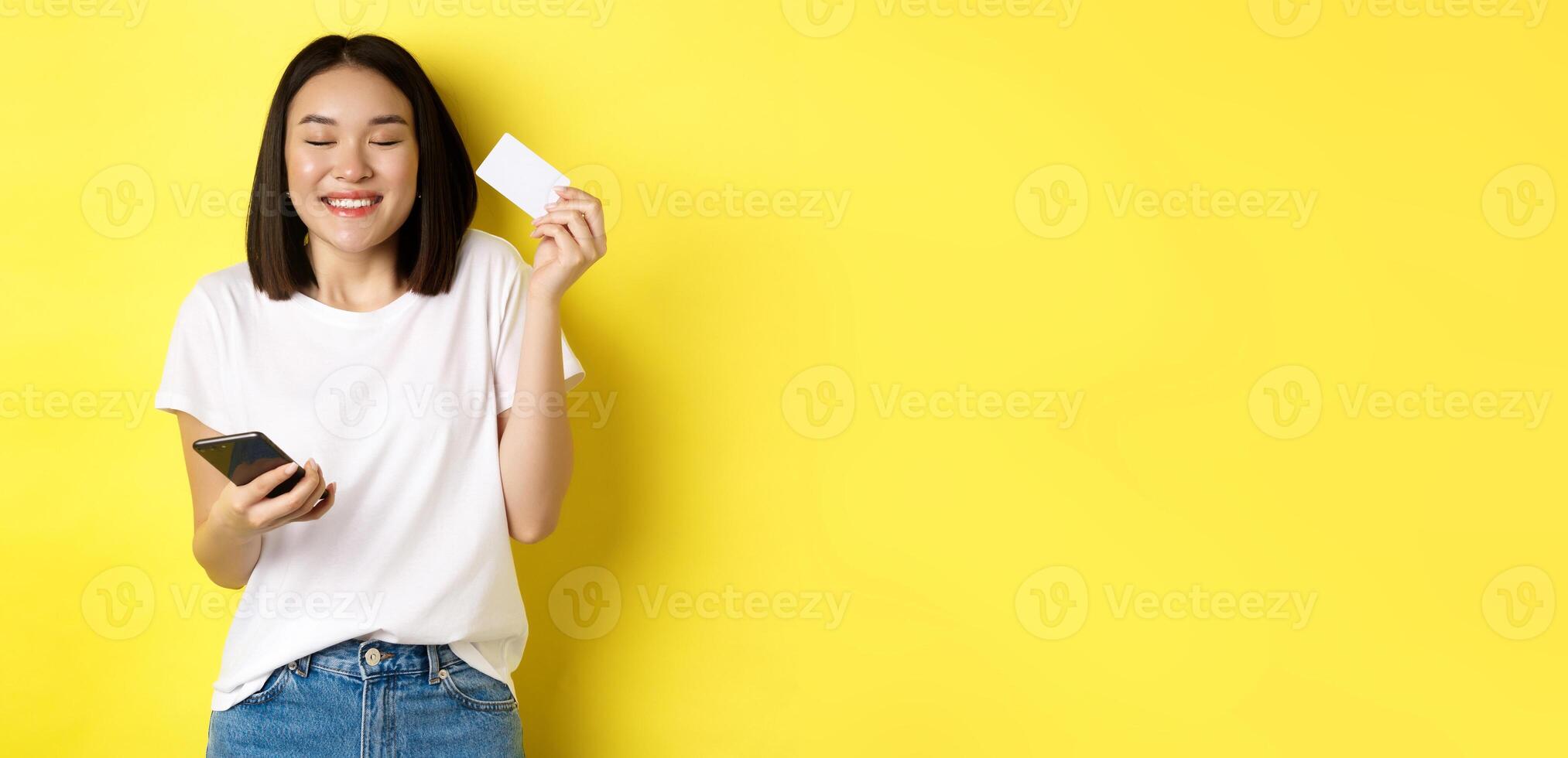 E-commerce and online shopping concept. Happy asian woman looking excited, buying something in internet, holding smartphone and showing plastic credit card, yellow background photo