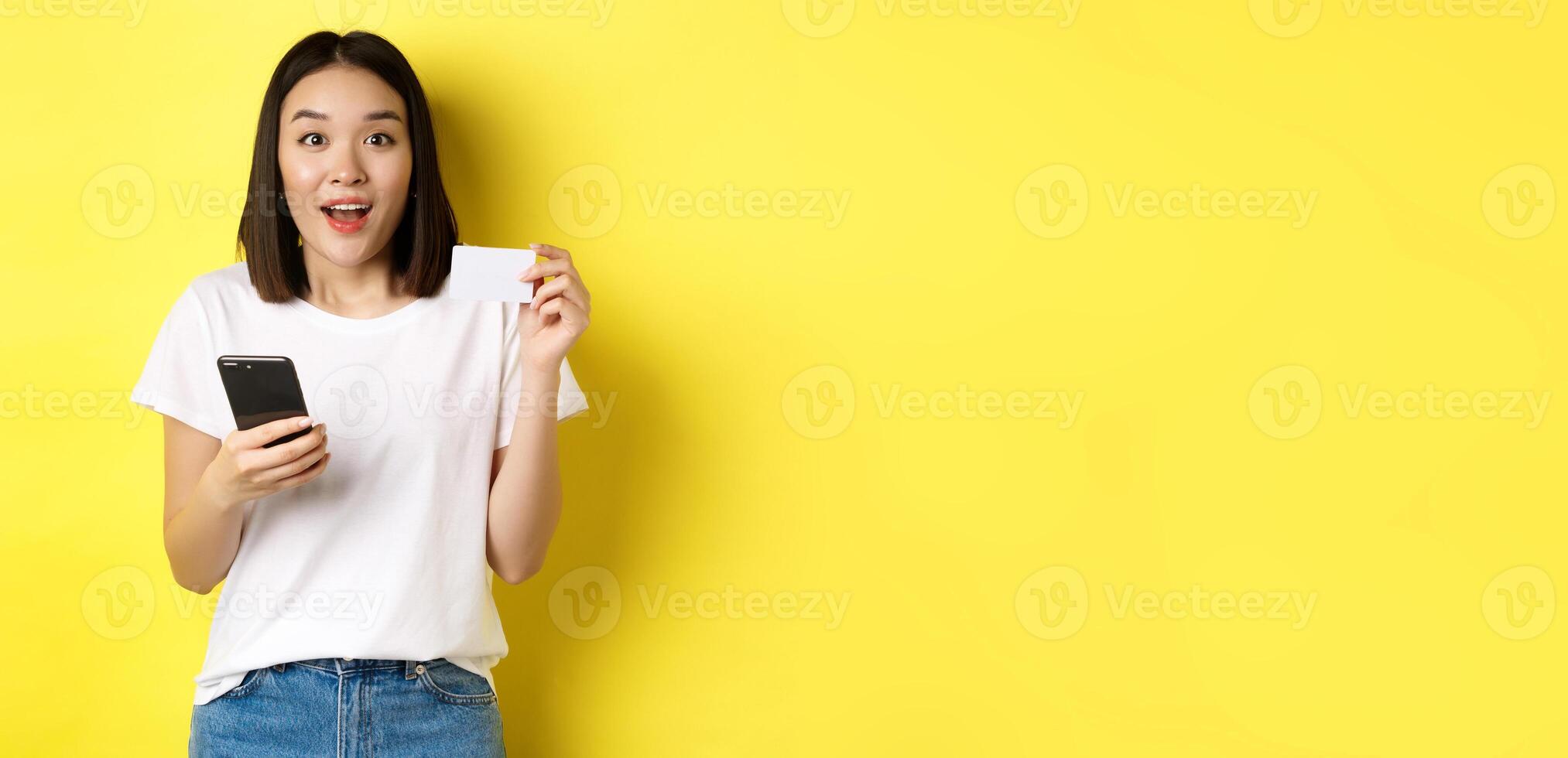E-commerce and online shopping concept. Excited asian woman order in internet, holding smartphone and plastic credit card, standing over yellow background photo