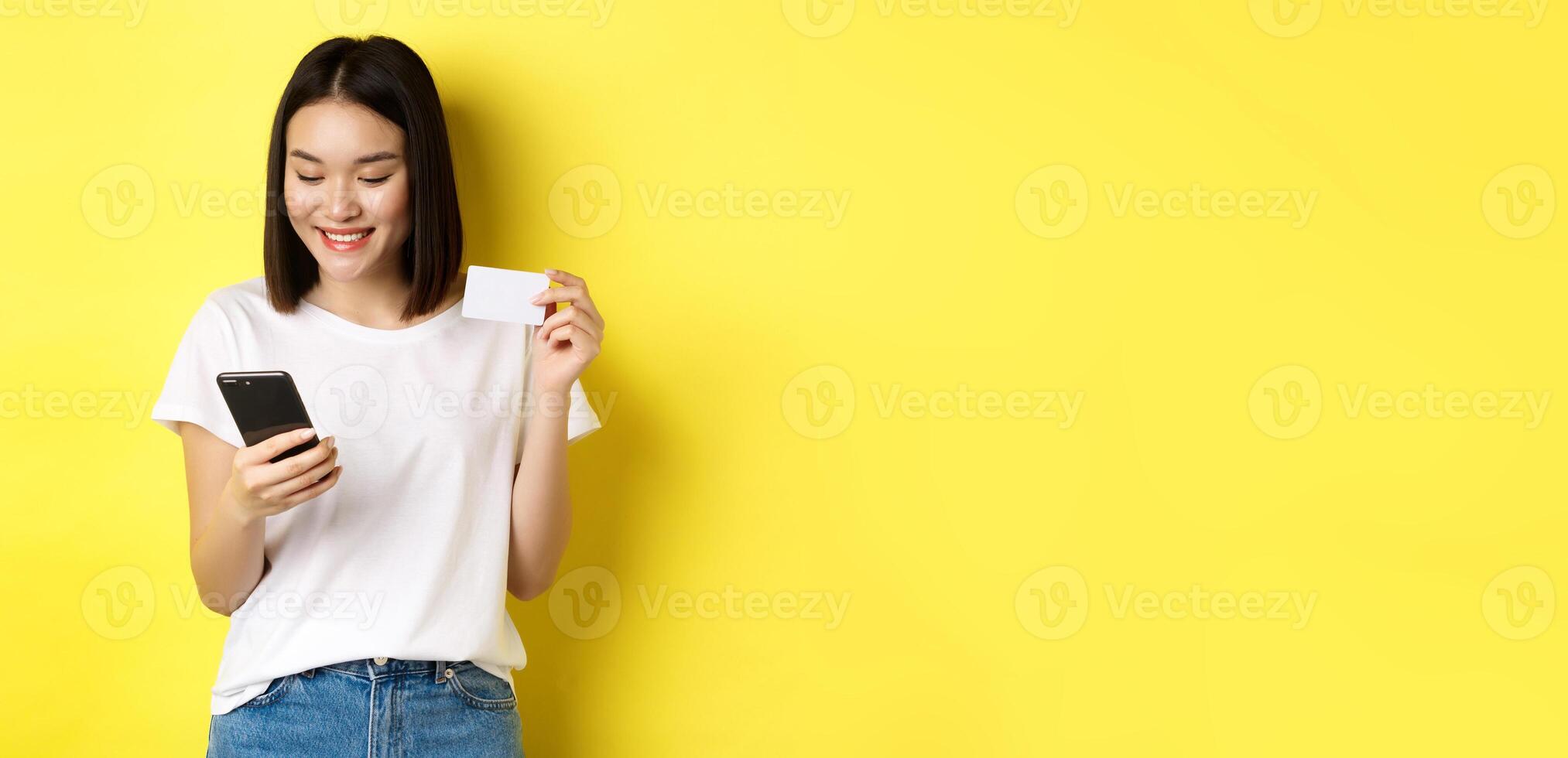 E-commerce and online shopping concept. Beautiful asian woman paying in internet, looking at smartphone screen and holding plastic credit card, yellow background photo