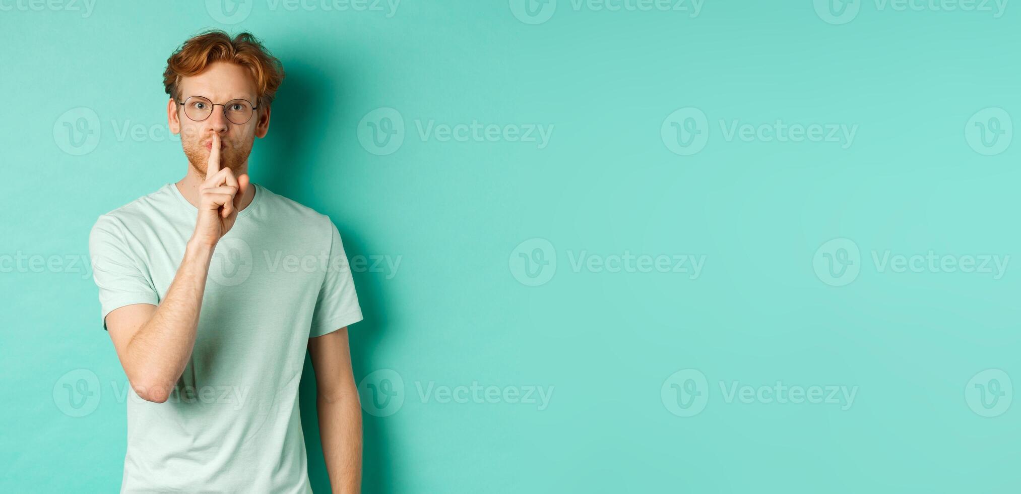 Serious redhead man hushing, tell to be quiet, making shush gesture and looking at camera, standing in t-shirt over turquoise background photo
