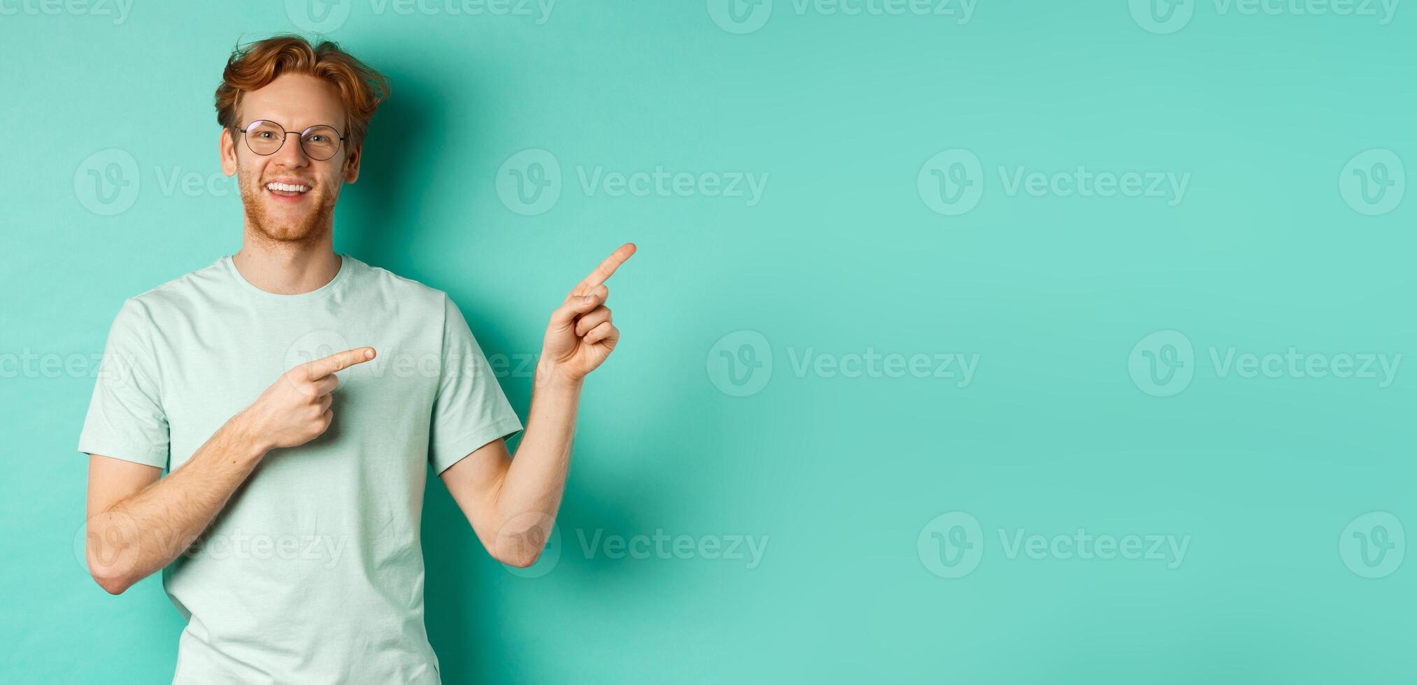 Handsome caucasian man with ginger hair, wearing glasses and t-shirt, pointing fingers right and smiling joyful, showing advertisement, standing over turquoise background photo