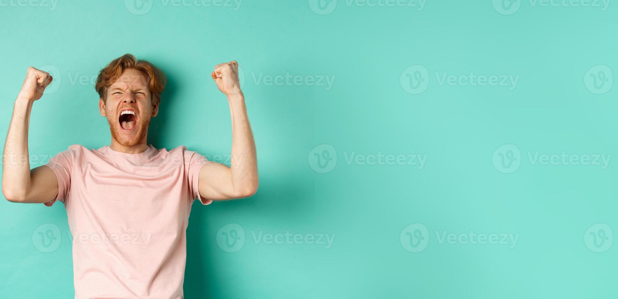 Young redhead man triumphing as watching sport game, making bet on winning team and rejoicing, screaming with joy and satisfaction, standing over turquoise background photo
