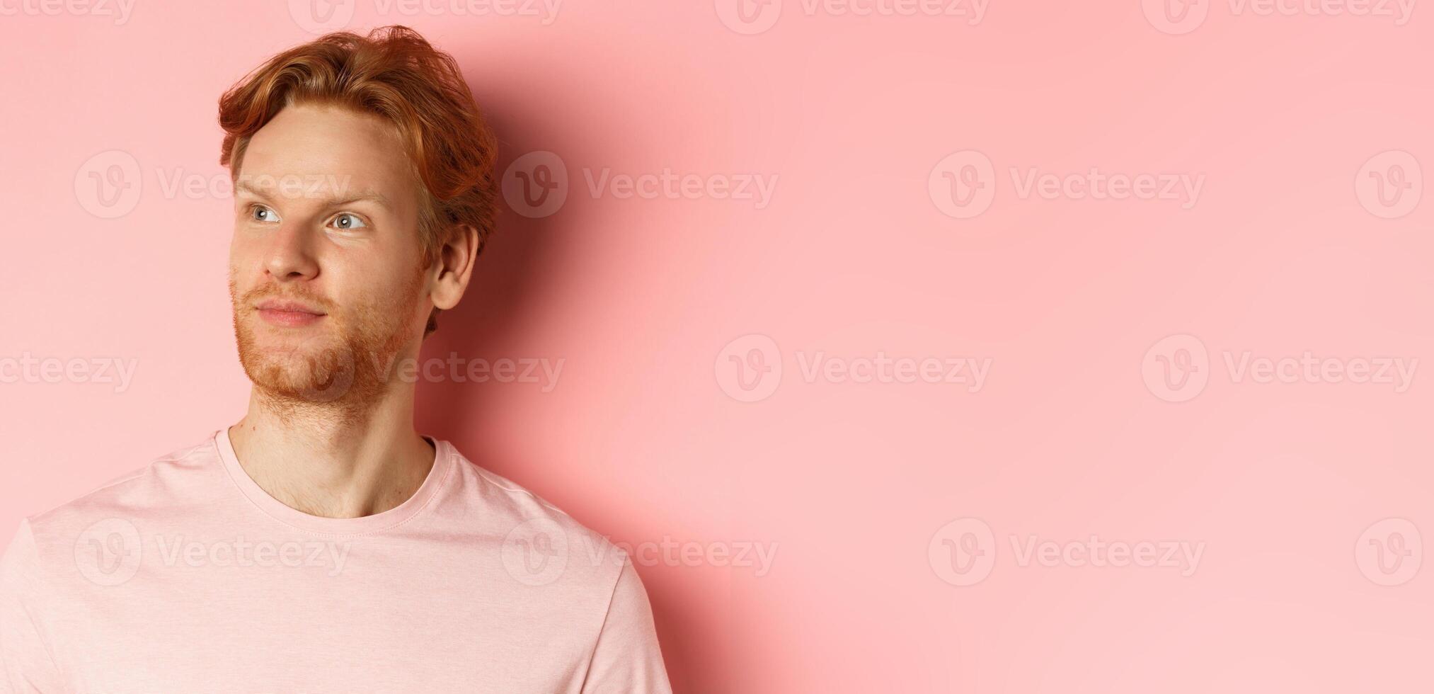Handsome european male model with red hair and beard, turn head and looking pleased at copy space on left side, standing over pink background photo