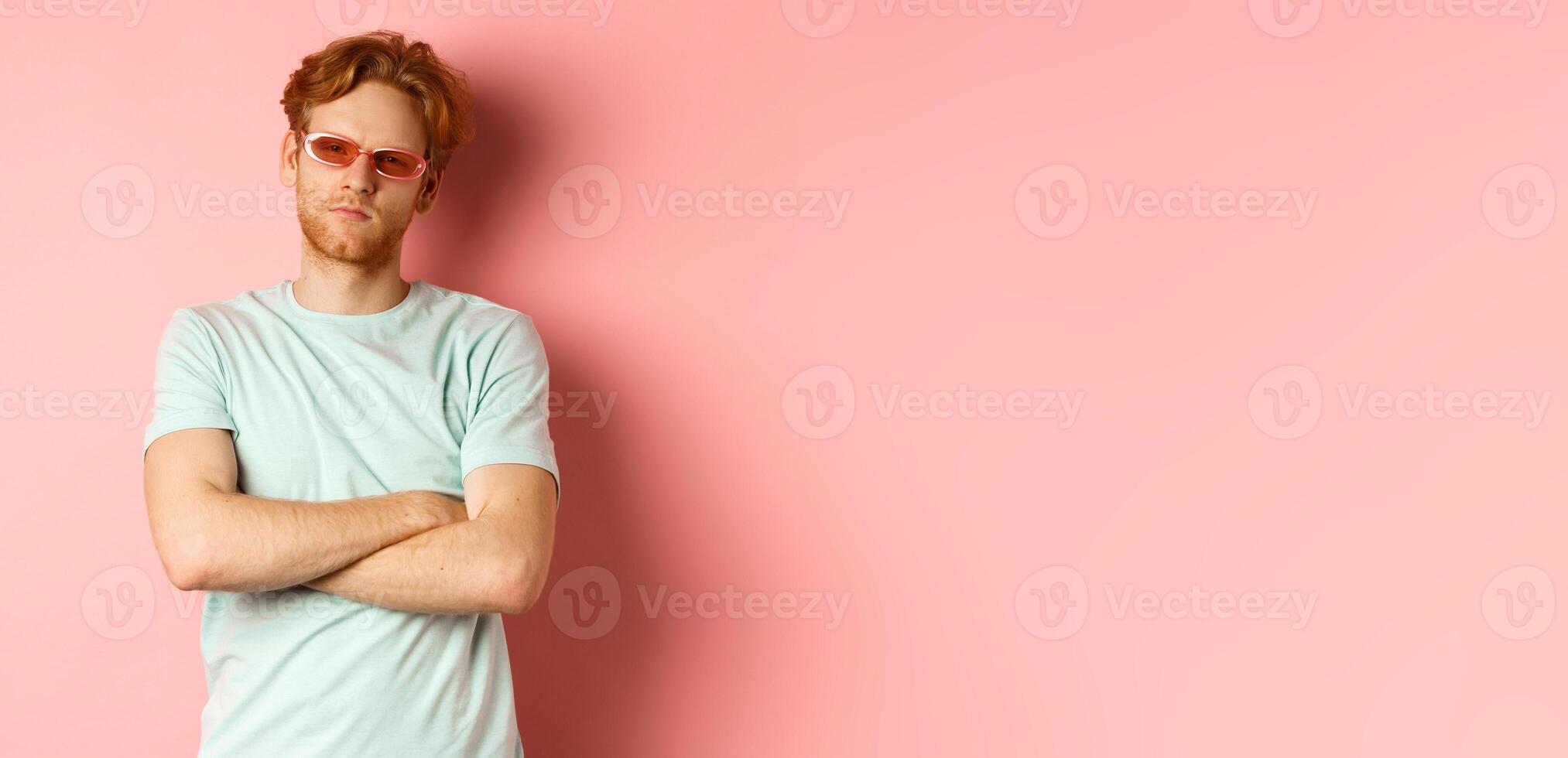 Tourism and vacation concept. Sassy redhead guy in sunglasses, cross arms on chest and looking serious at camera, standing over pink background photo