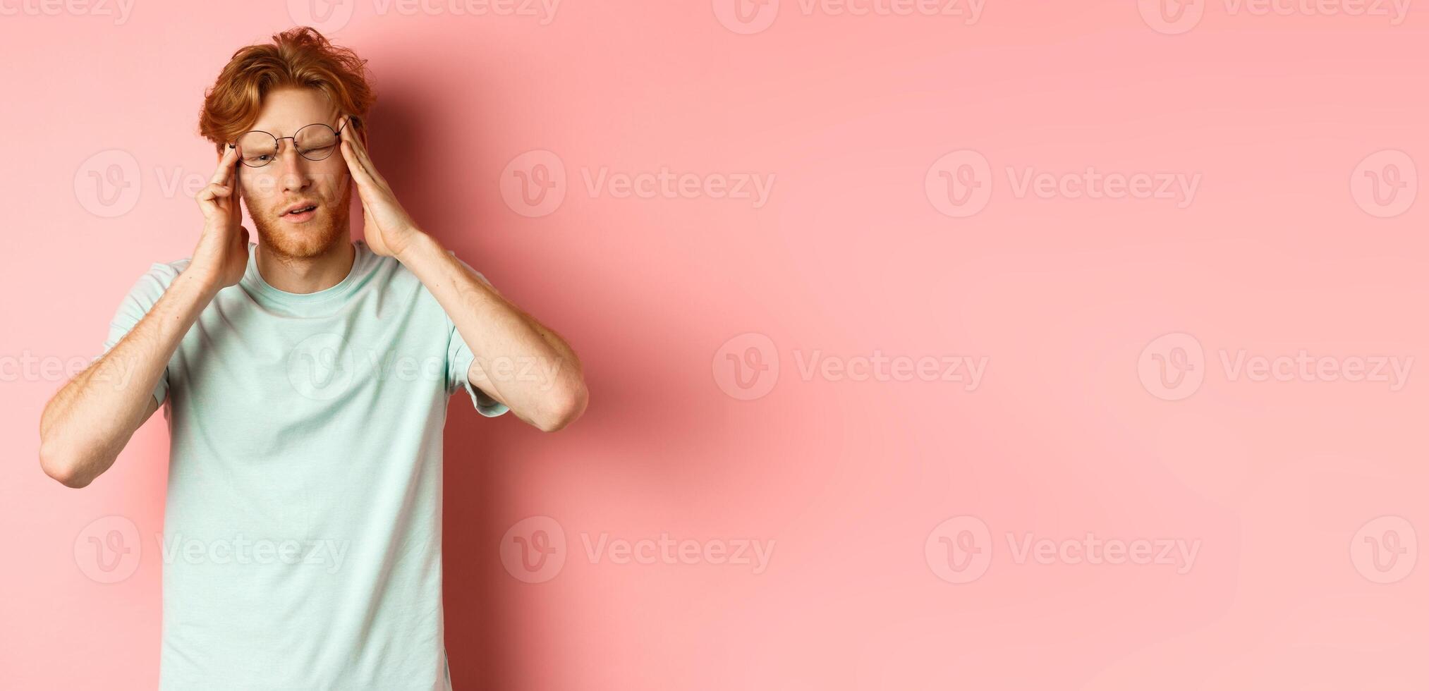 Portrait of redhead man in crooked glasses touching head and feeling dizzy or nauseous, having hangover or headache, standing over pink background photo