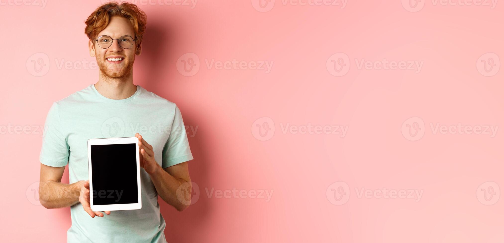 Funny redhead guy in glasses demonstrate digital tablet screen, smiling at camera, standing over pink background photo
