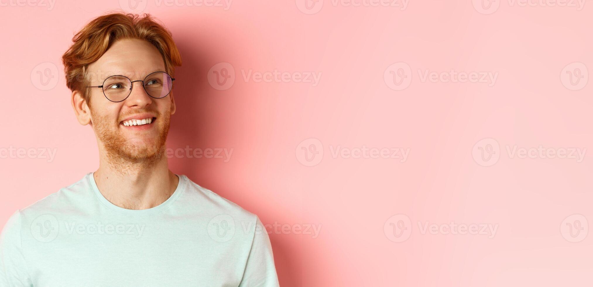 Face of handsome redhead man in glasses smiling pleased, looking at upper right corner, standing over pink background photo