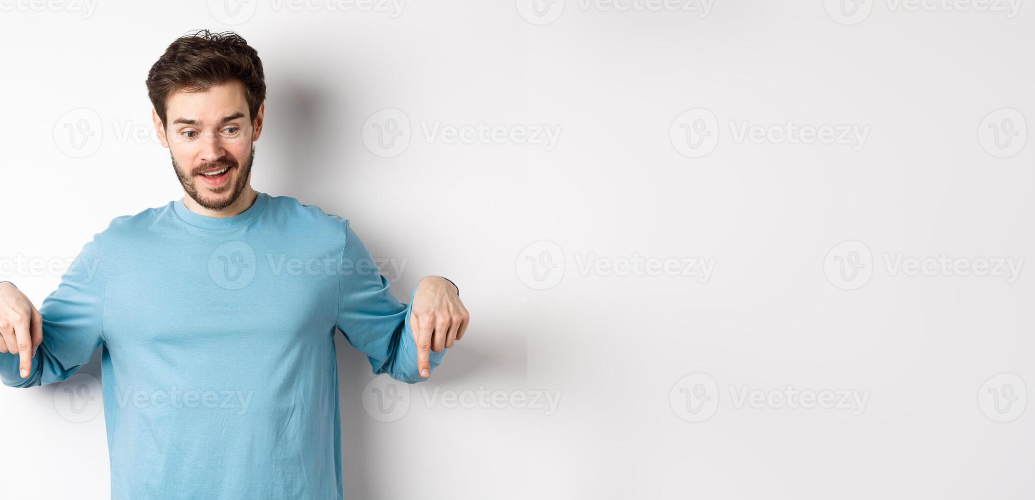 Handsome caucasian man pointing down with impressed and happy face, smiling and saying wow, standing on white background photo